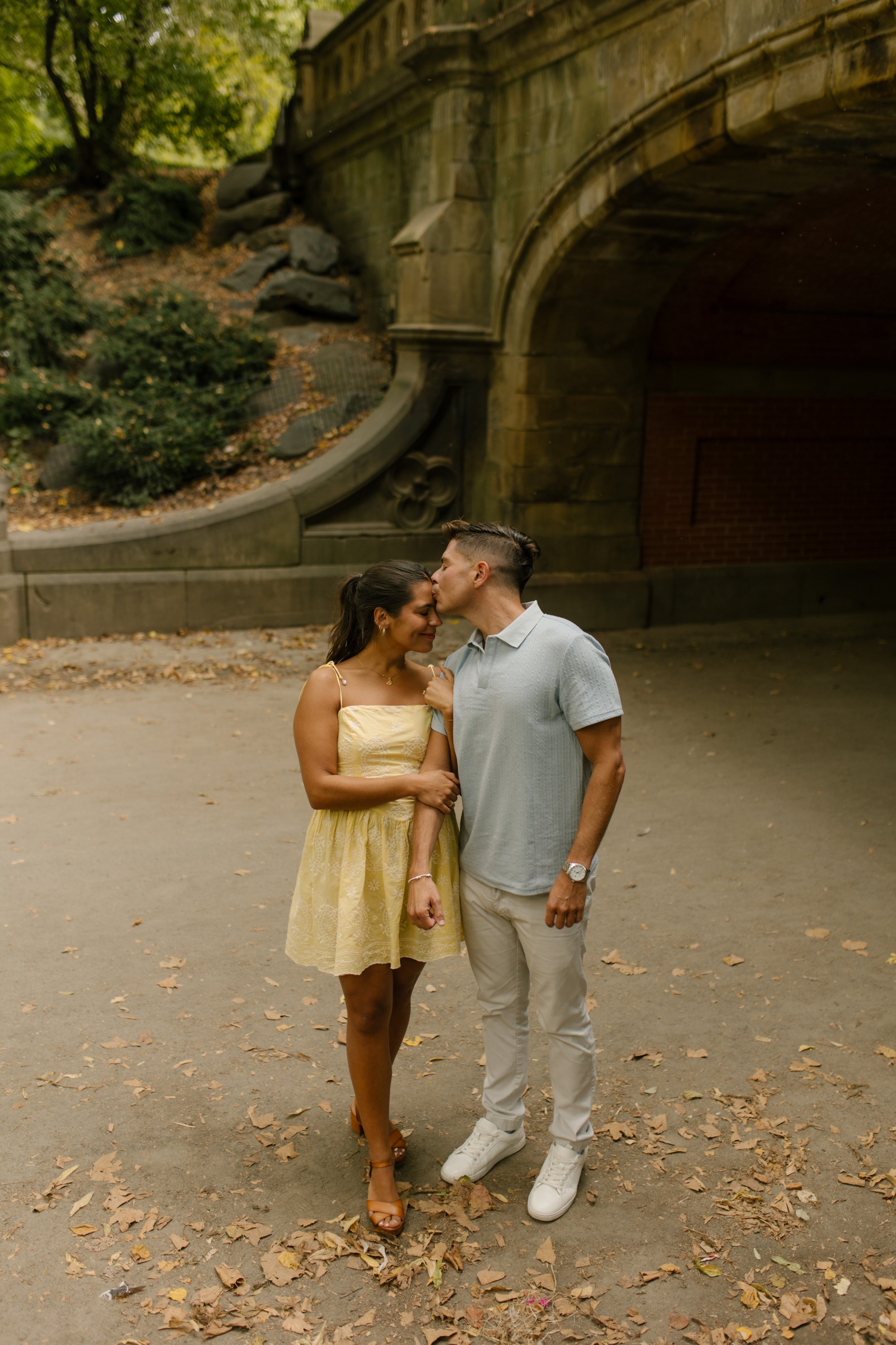 Chris kisses Sienna on the cheek as they stand together beneath a stone archway in Central Park, surrounded by fallen leaves.