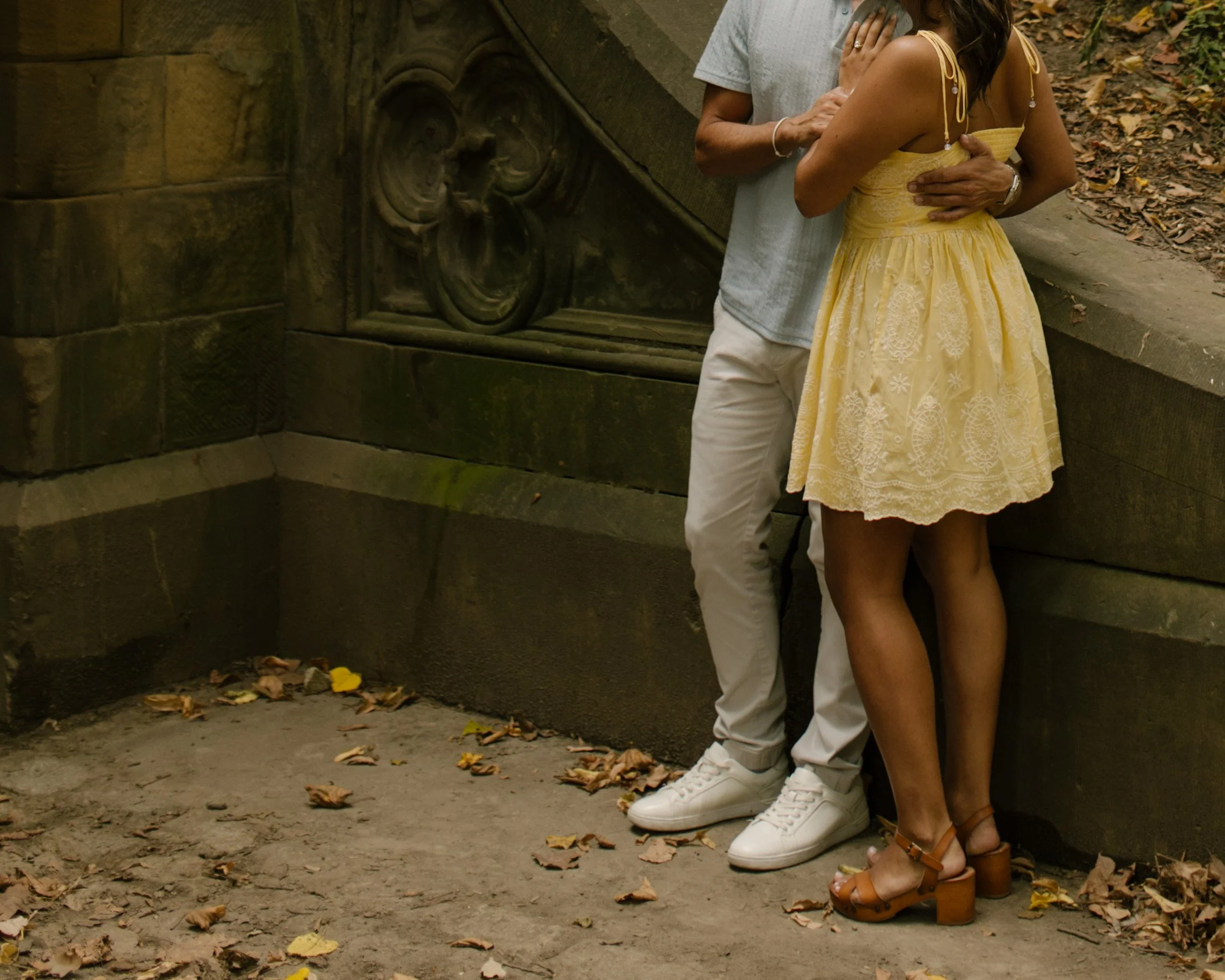 Full-length photograph of Chris and Sienna embracing beside a curved stone wall, showing Sienna's yellow floral dress and brown heels.