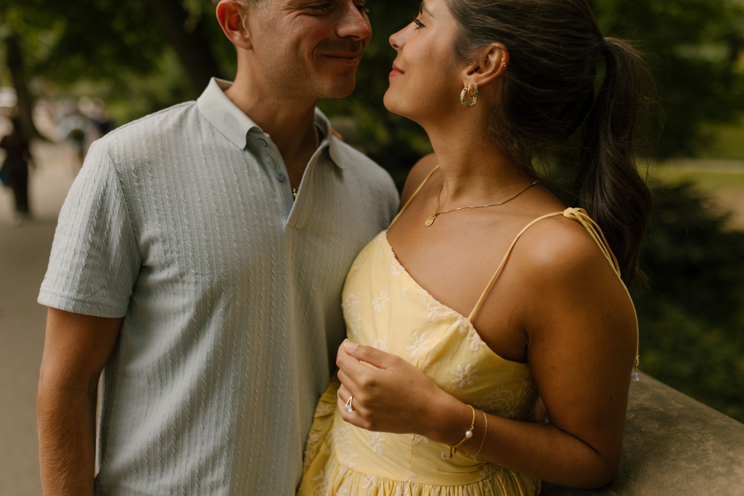 Sienna gazes up at Chris while standing close beside a stone railing during their new york proposal portraits in Central Park.