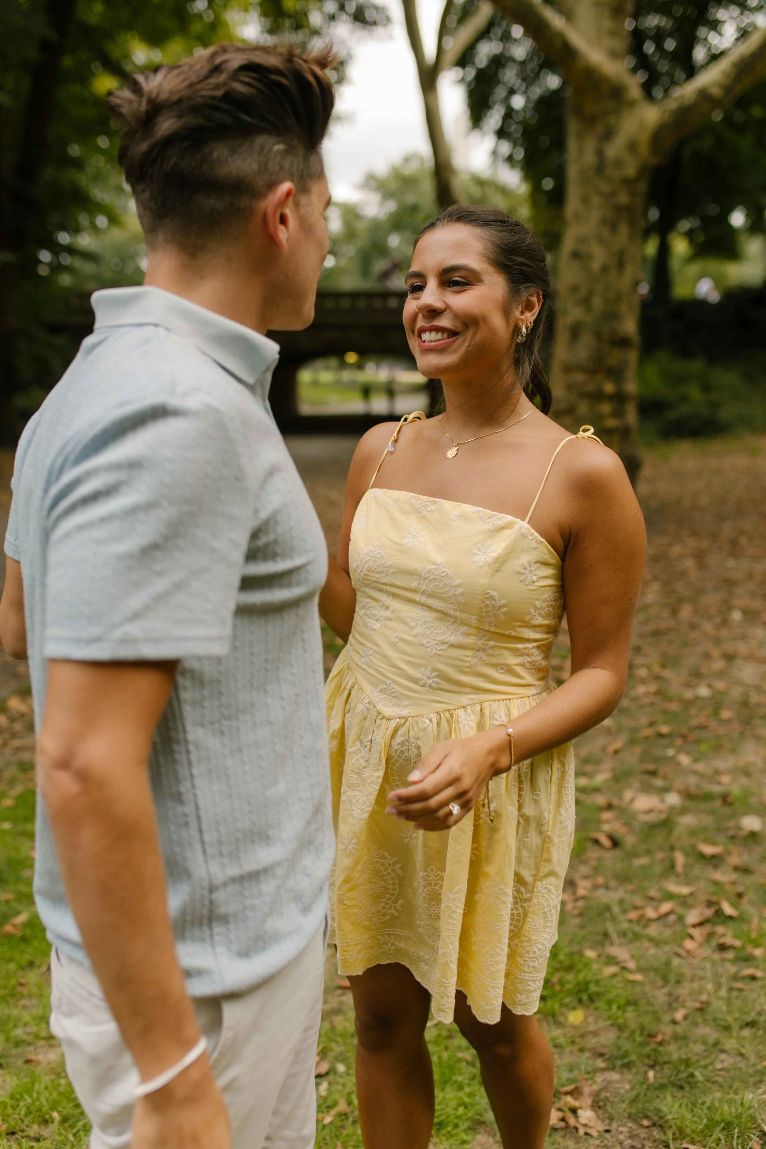 Chris and Sienna share a laugh on a grassy pathway surrounded by trees, with Sienna wearing a pale yellow dress and Chris in a light blue polo.