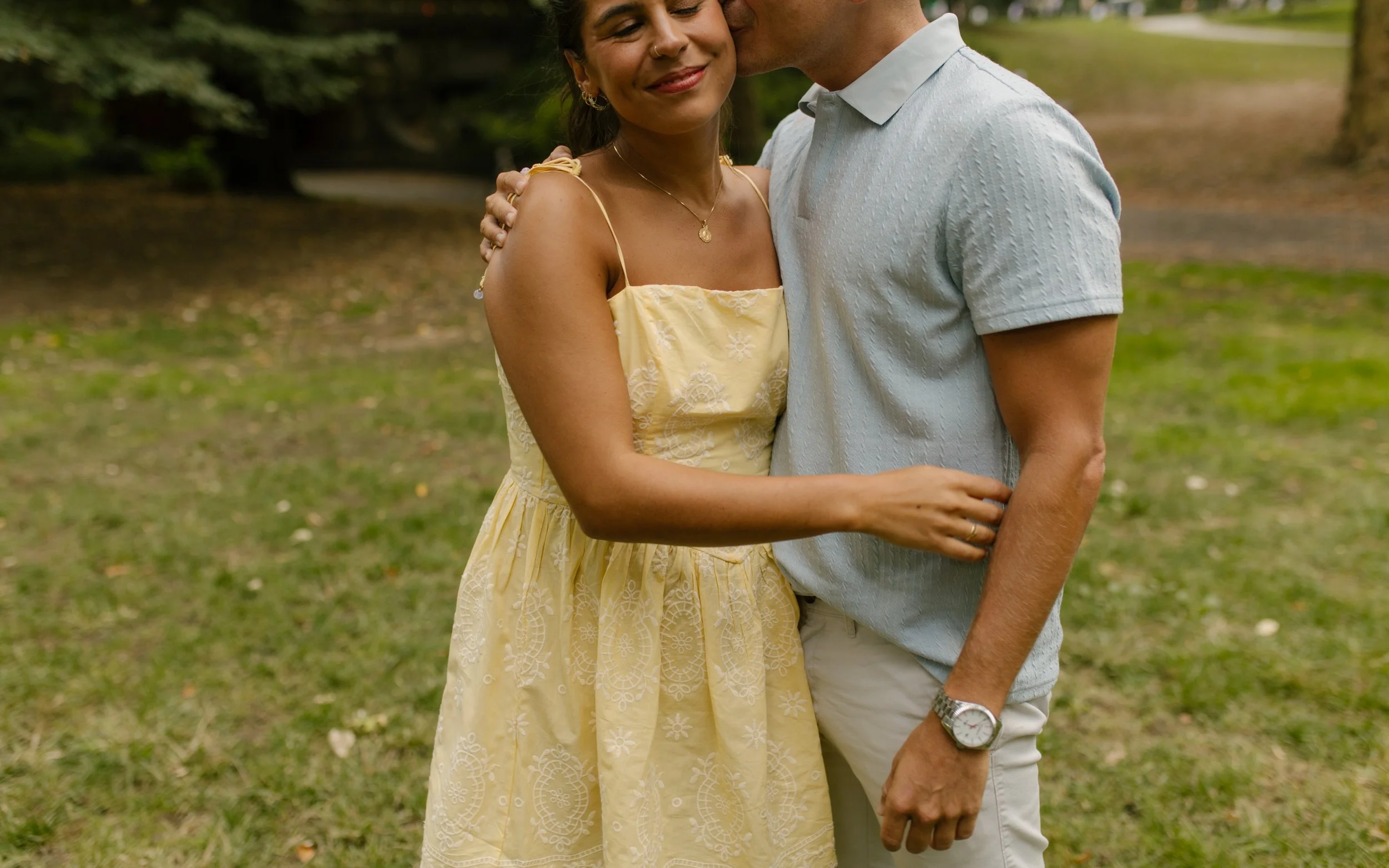 Chris kisses Sienna on the temple as they embrace on a grassy lawn, with her yellow floral dress and his light blue polo visible.