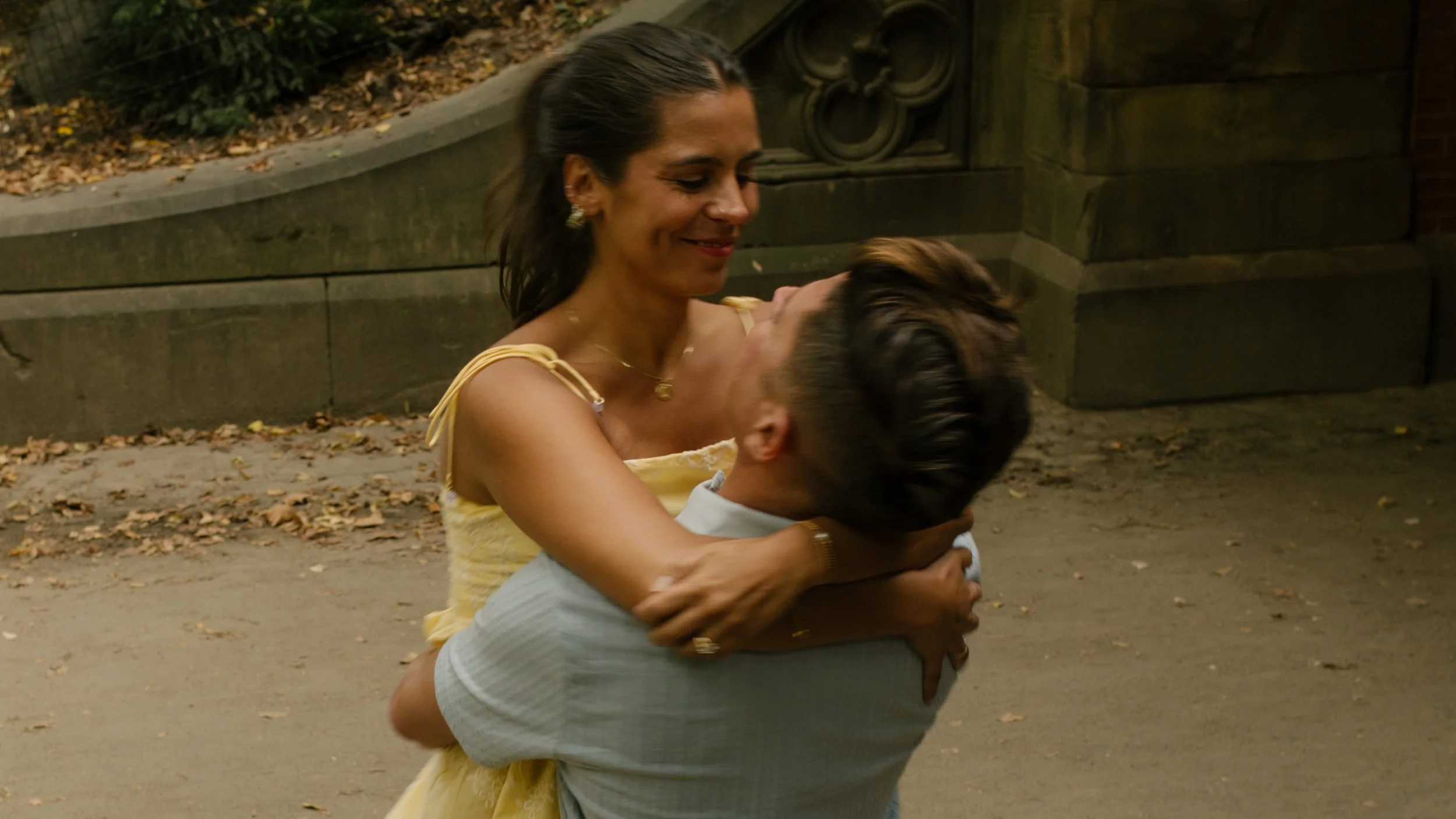 Sienna smiles down at Chris as he holds her in his arms near a historic Central Park stone bridge, celebrating their romantic new york proposal