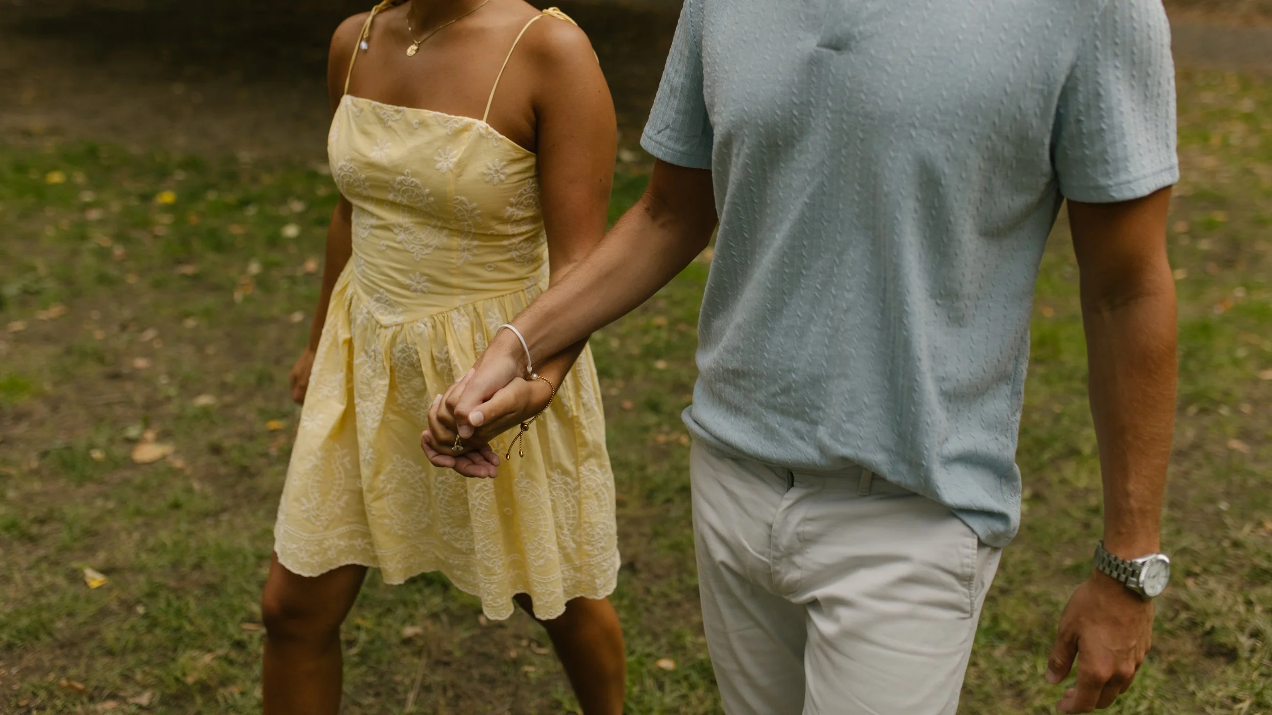 Close-up of Chris and Sienna walking hand in hand through Central Park, with Sienna's yellow floral dress and Chris's light blue polo visible.