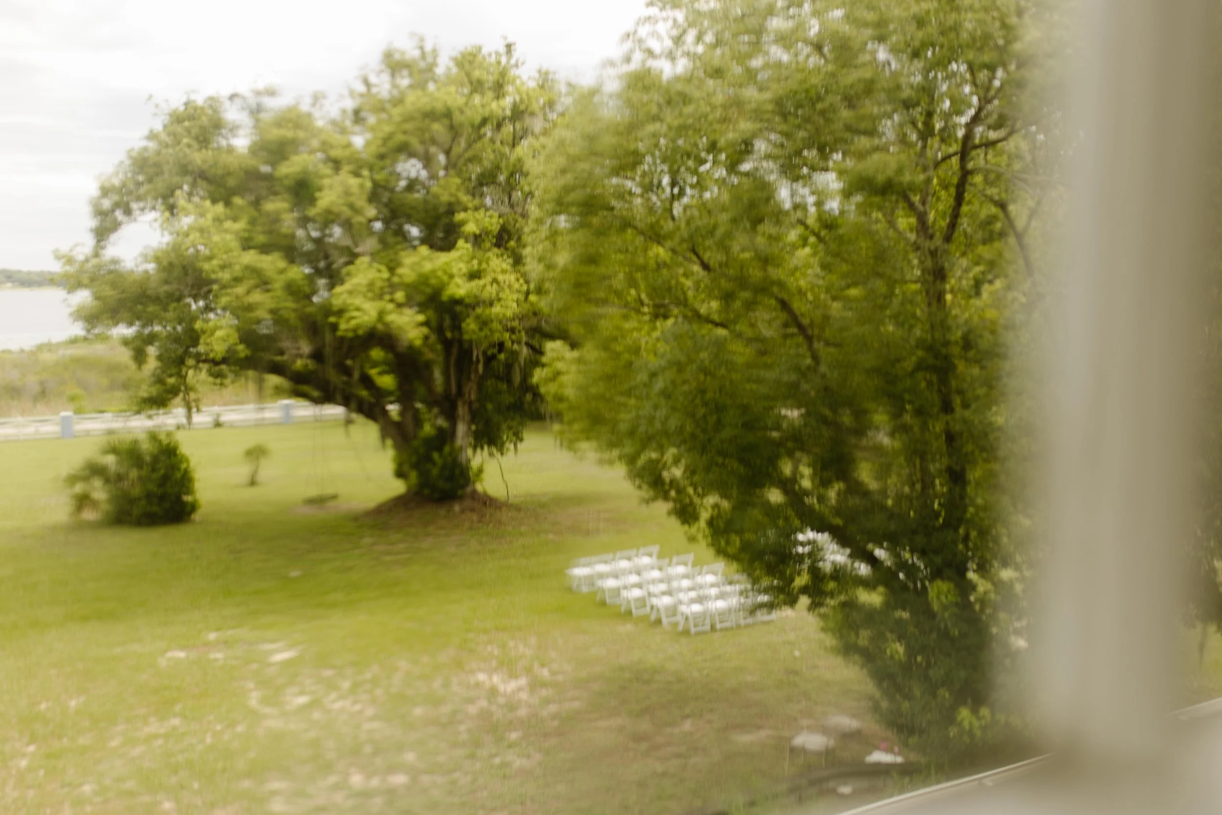 Blurred view through a window of the wedding weekend ceremony setup beneath lush trees by the water.