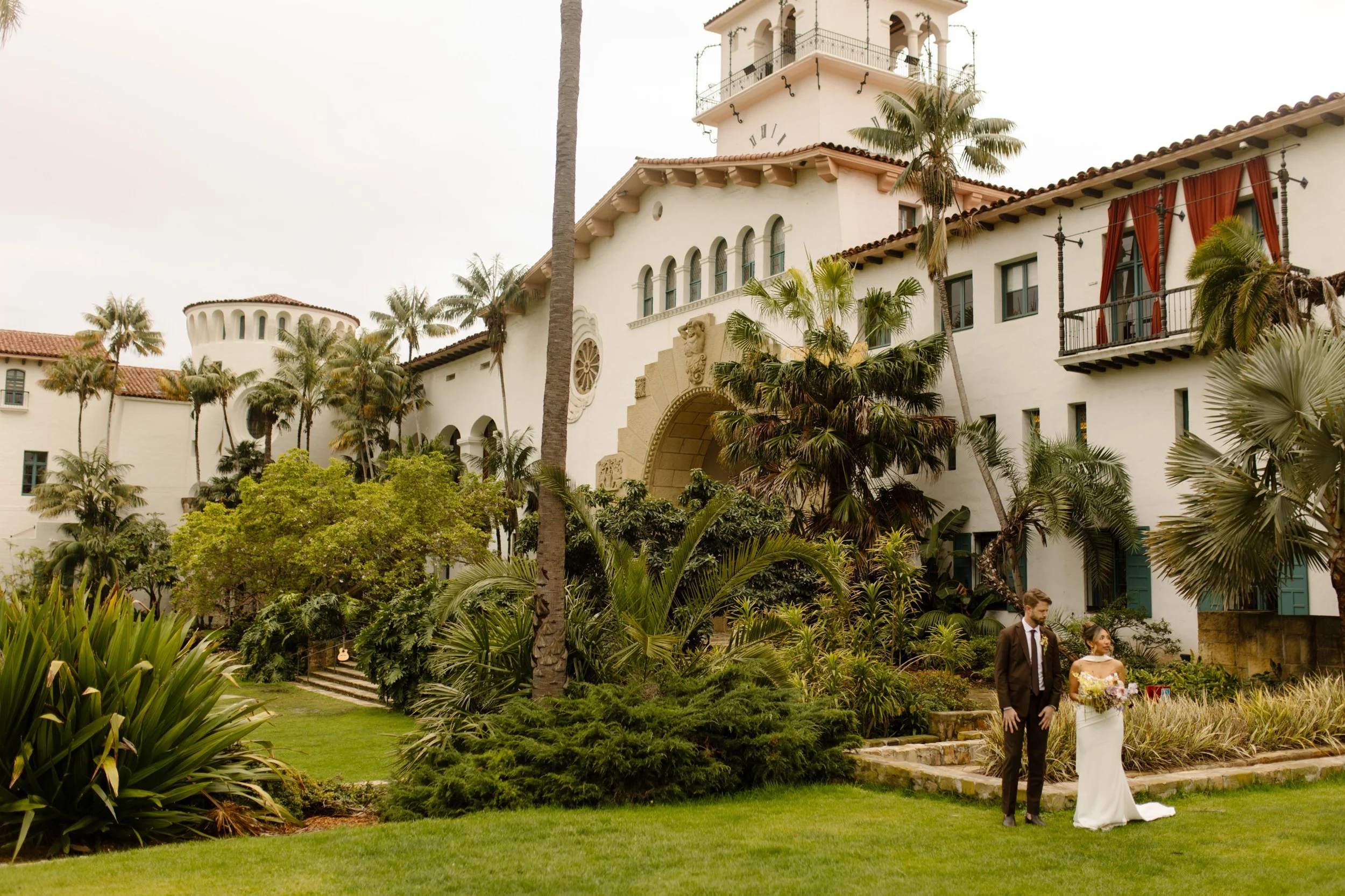 Wide shot of the couple walking the lawn, framed by palm trees and the wedding Santa Barbara Courthouse.