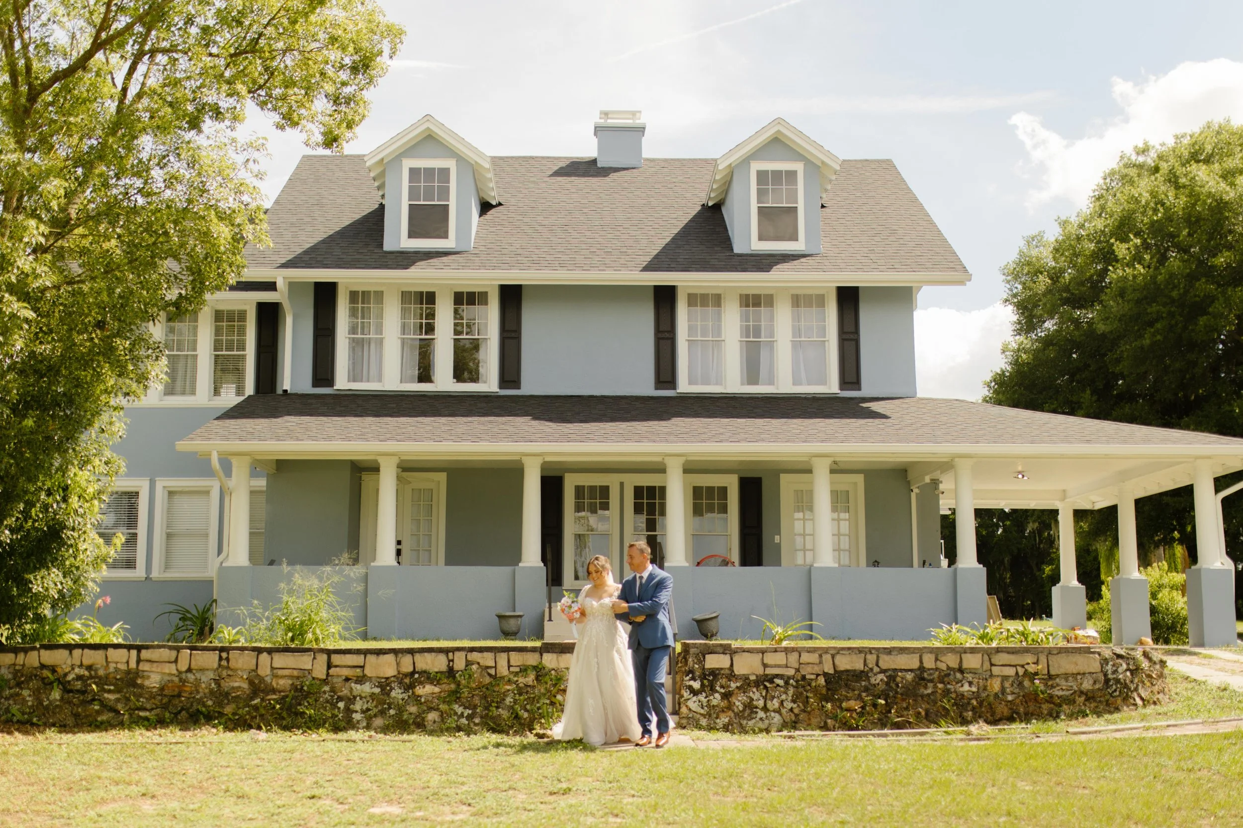 Bride walking arm-in-arm with her father down the lawn of a charming blue house, ready to begin her wedding weekend ceremony.