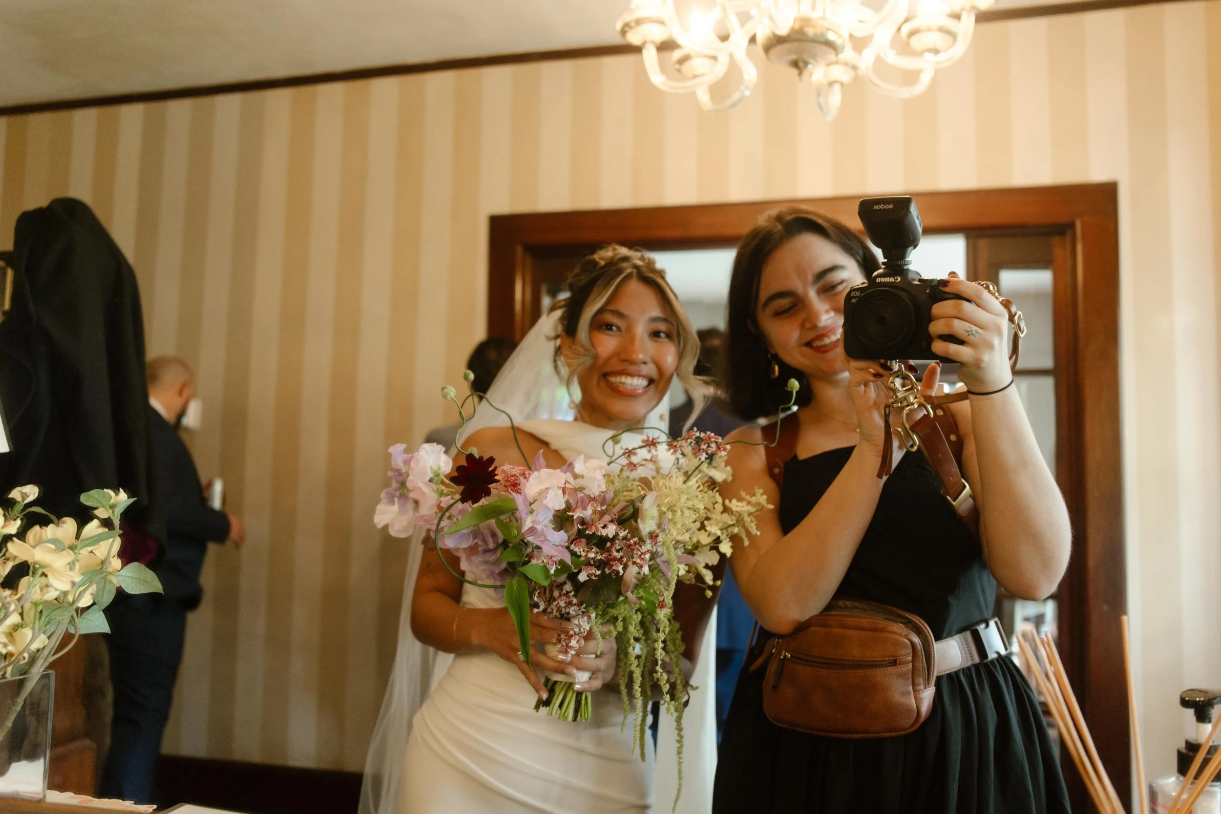 A joyful bride holds her bouquet and smiles beside her photographer, capturing a moment of pre-ceremony excitement.