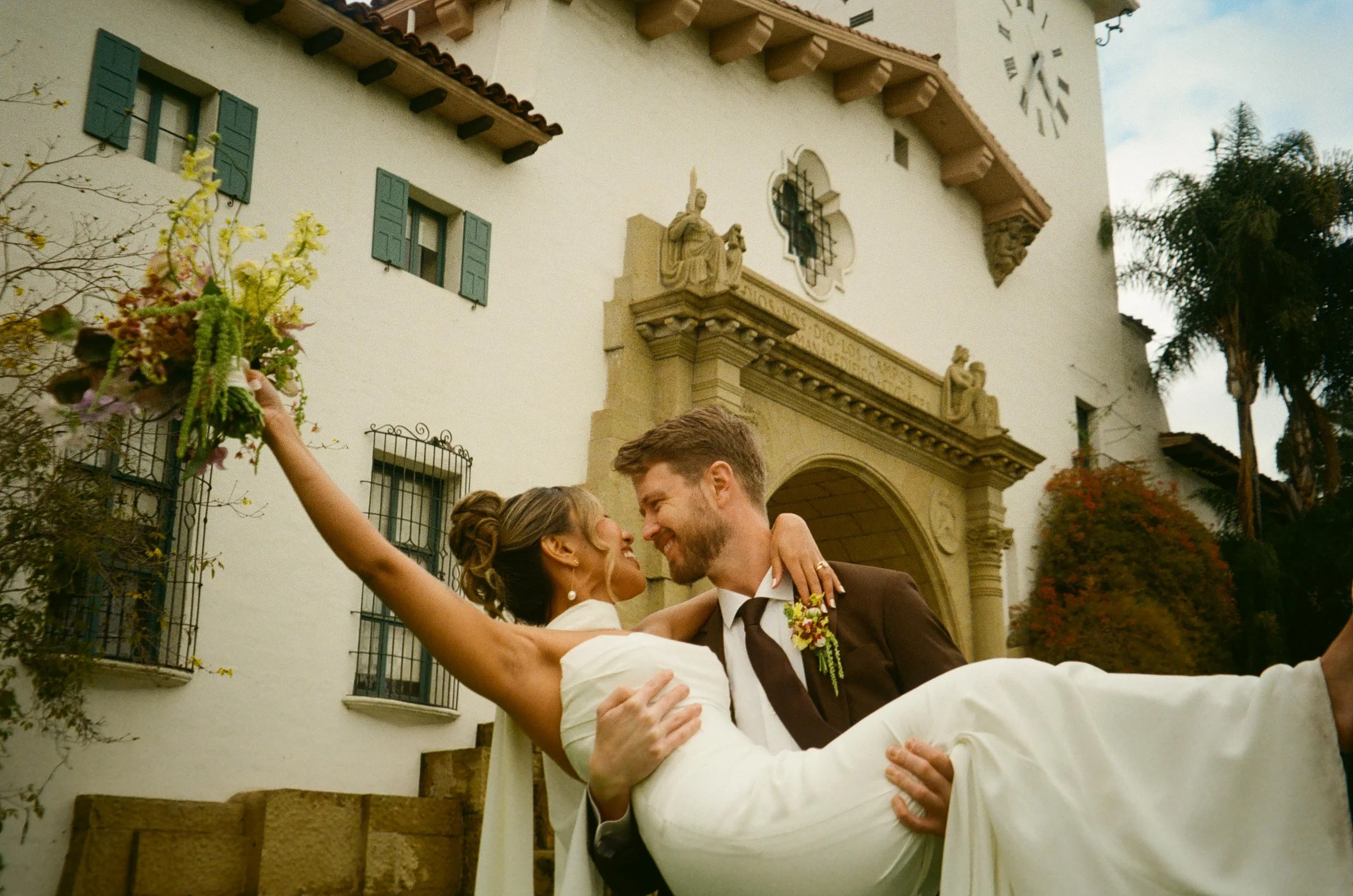 The groom lifts the bride in front of the clock tower, bouquet raised high—pure joy at their wedding Santa Barbara Courthouse.