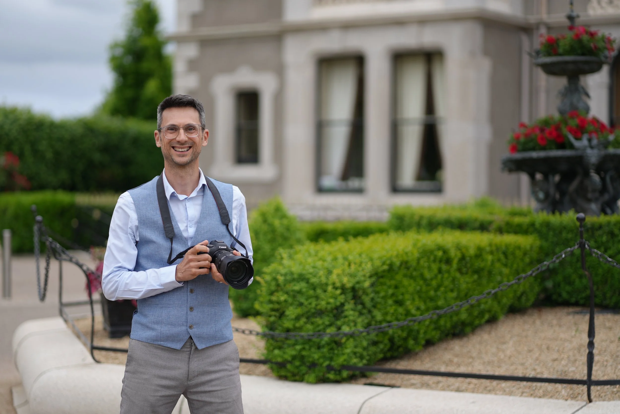 A man with glasses smiling and holding a camera in front of a large building with windows and a garden with red flowers.