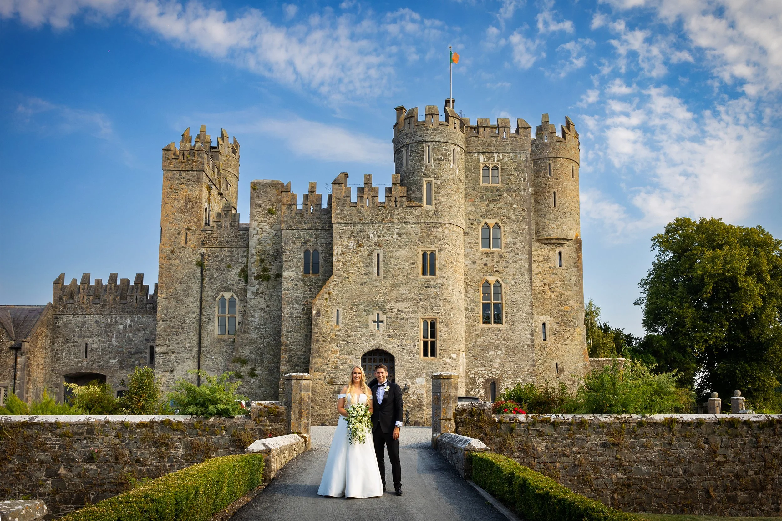 A bride and groom standing in front of a large stone castle with turrets, Irish flag, and a clear blue sky. The bride is wearing a white wedding dress and holding a bouquet, while the groom is in a black tuxedo.