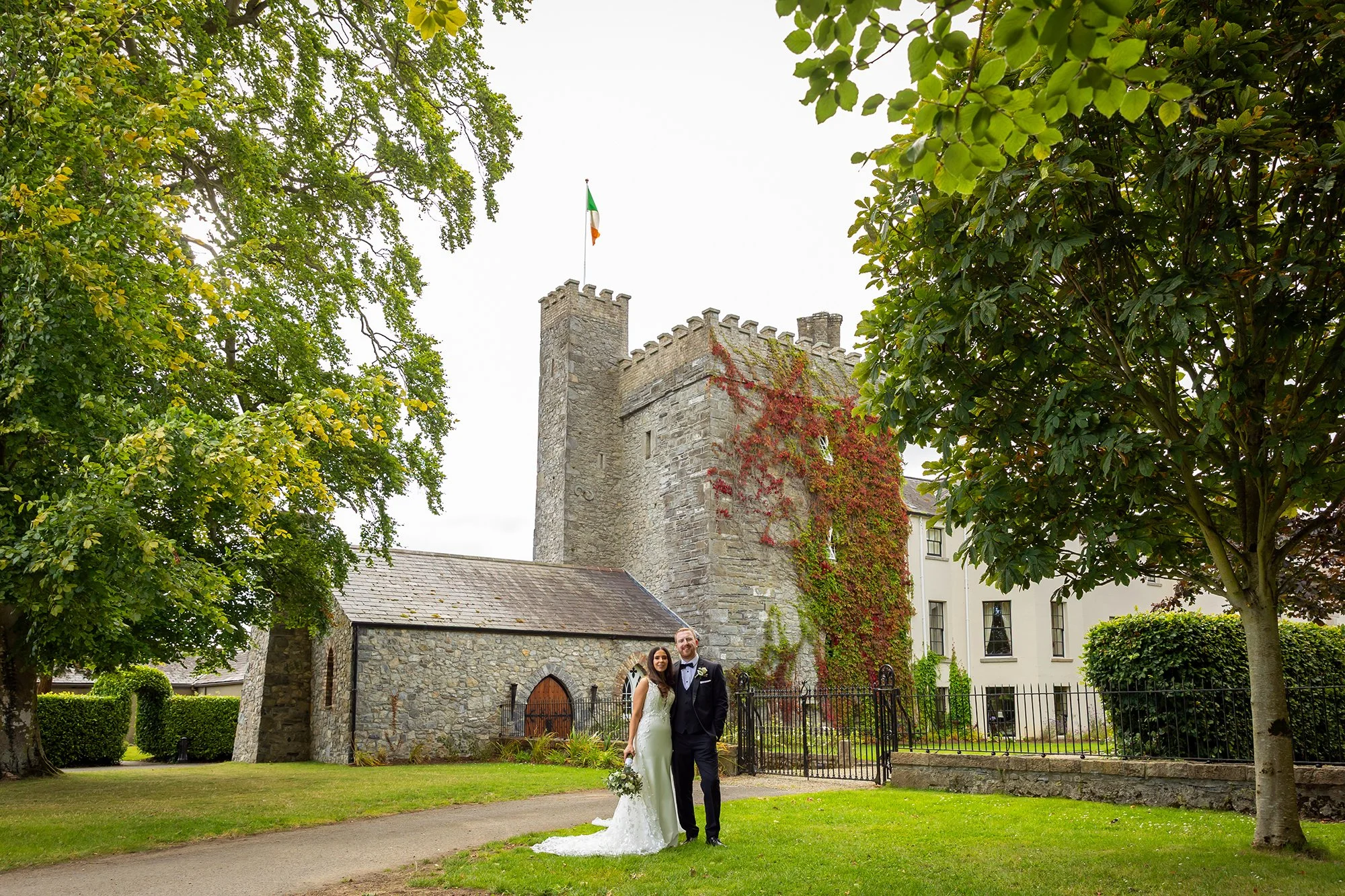 A bride and groom standing together outside a historic stone church with a flag on a tower, surrounded by trees and greenery.