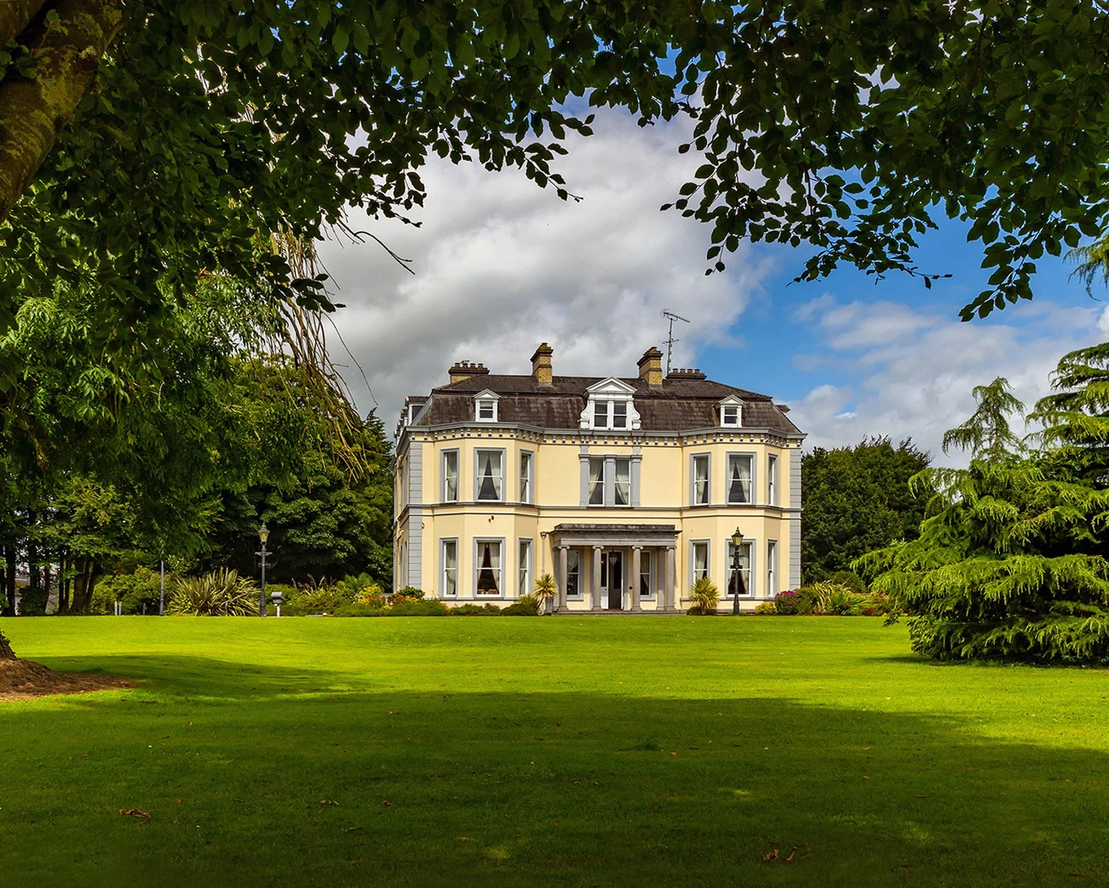 A large, elegant yellow house with white trim, surrounded by a lush green lawn and tall trees, under a cloudy sky.