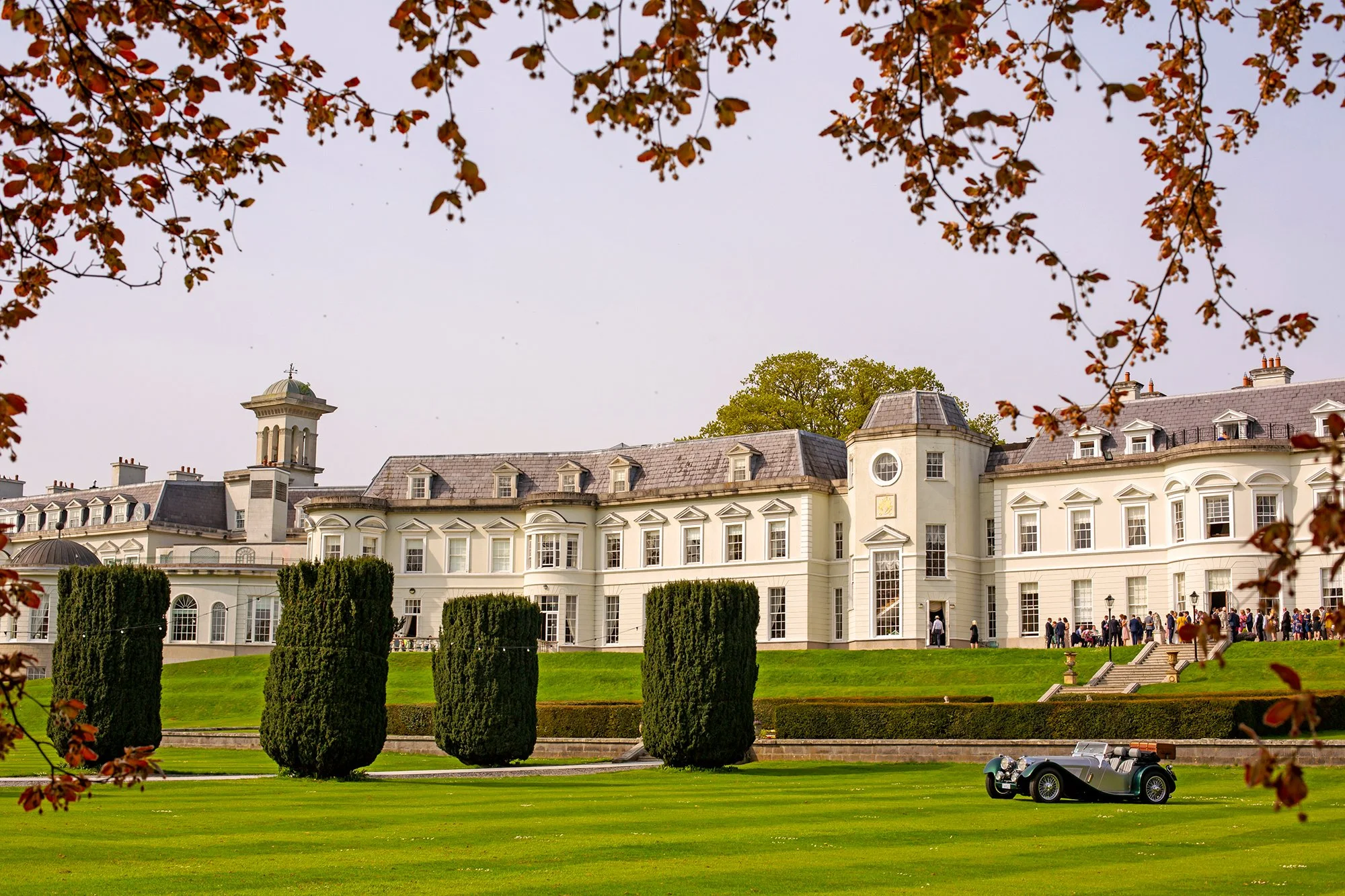 A large white historic mansion with a golf cart parked on the grass in front. The mansion has chimneys, dormer windows, and a cupola with a cross. There are trees, some with autumn-colored leaves, framing the top of the image. People are gathering near the entrance of the mansion.