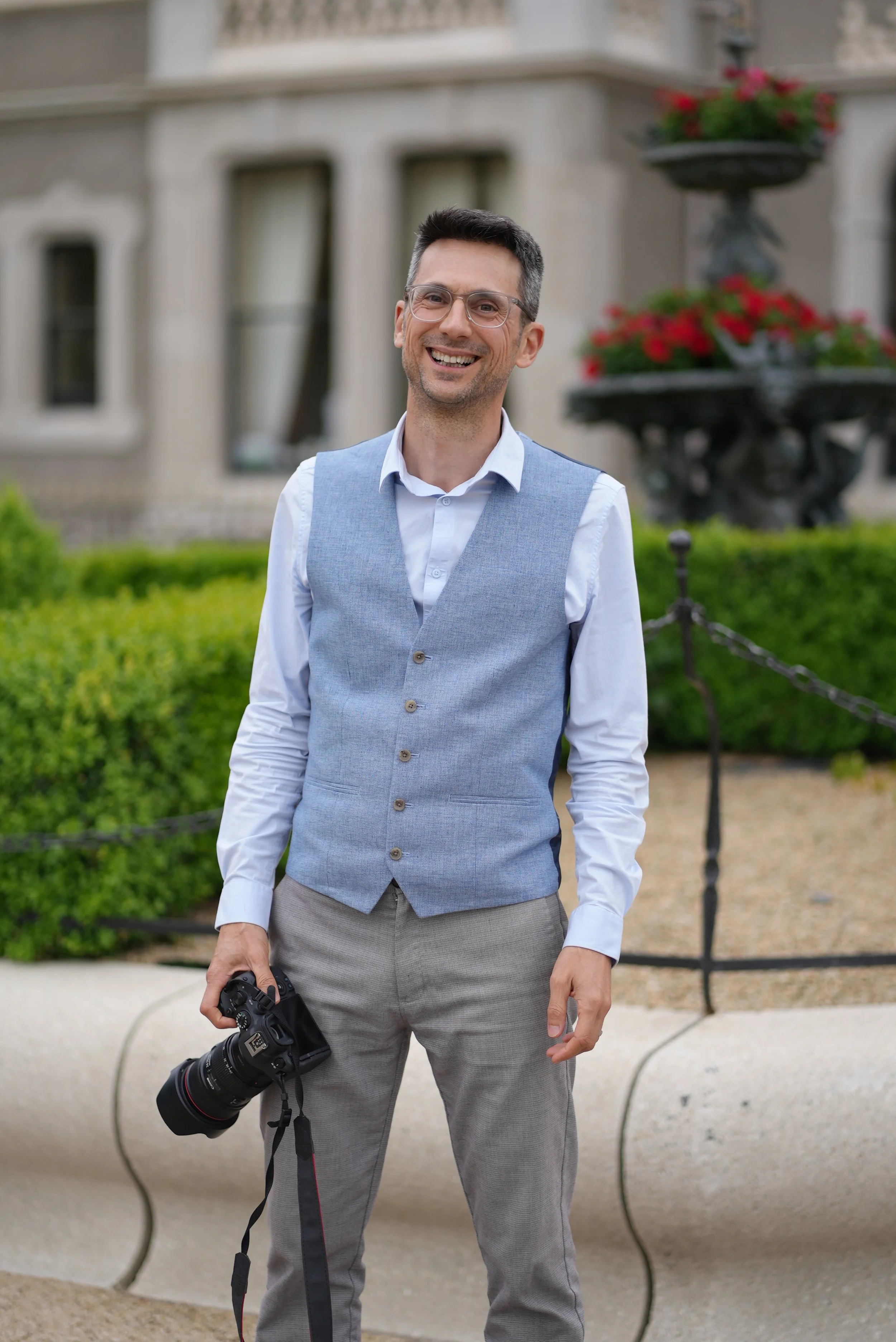 A man with glasses, wearing a white shirt and light gray vest, smiling while holding a camera, standing outdoors in front of a building with windows and a flowered planter.