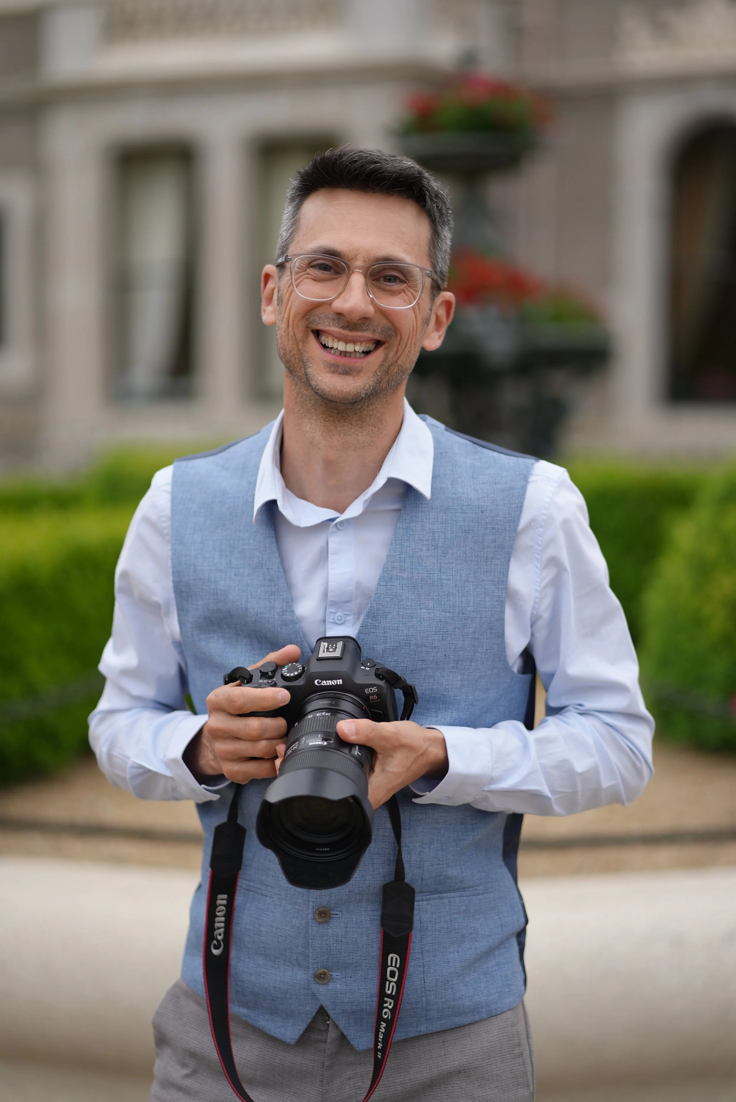 A smiling man wearing glasses, a white shirt, and a light blue vest holding a professional Canon DSLR camera outdoors in front of a blurred background of a building and greenery.