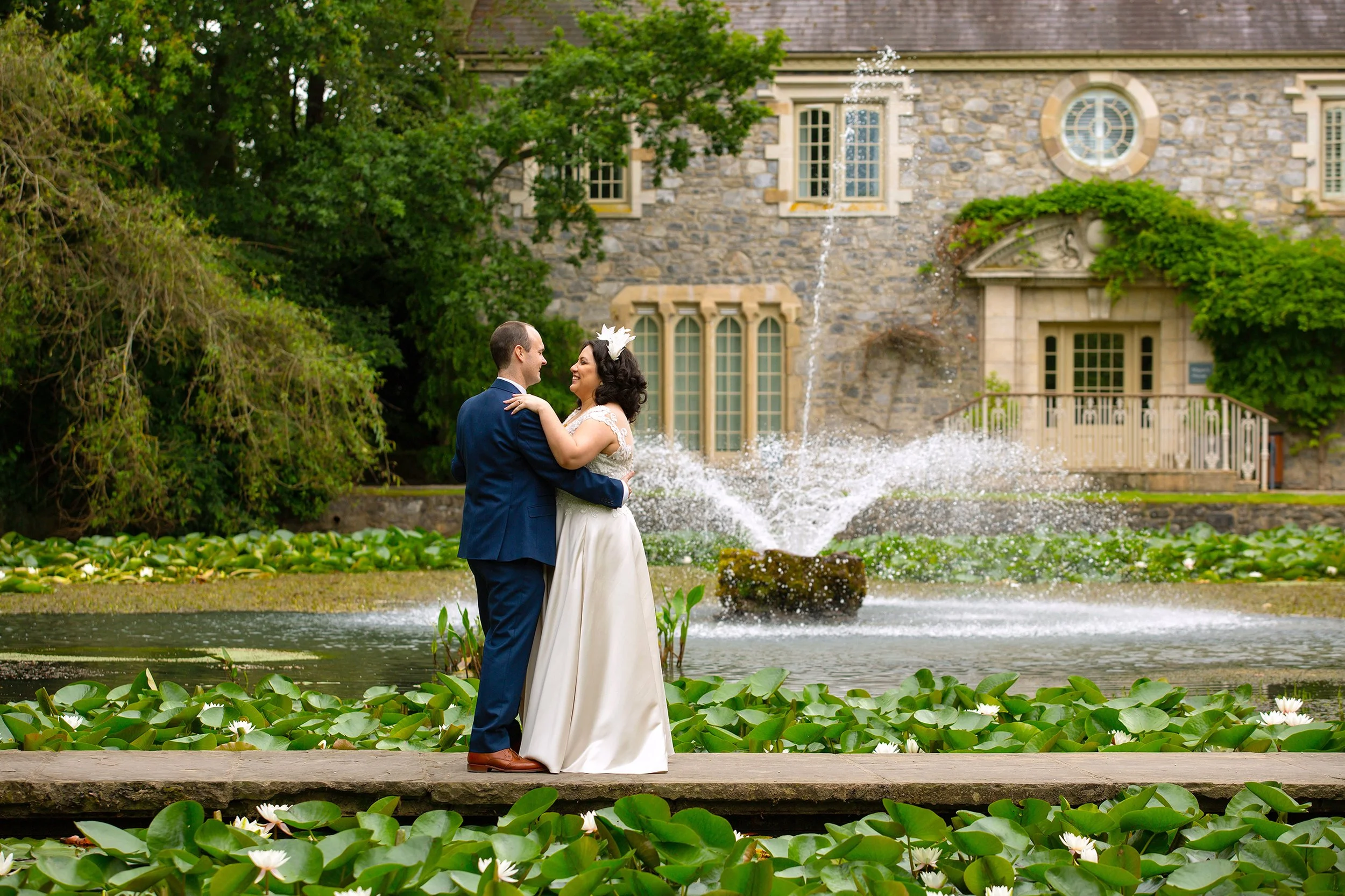 A bride and groom in wedding attire sharing a dance near a fountain and a stone building with arched windows and lush greenery.