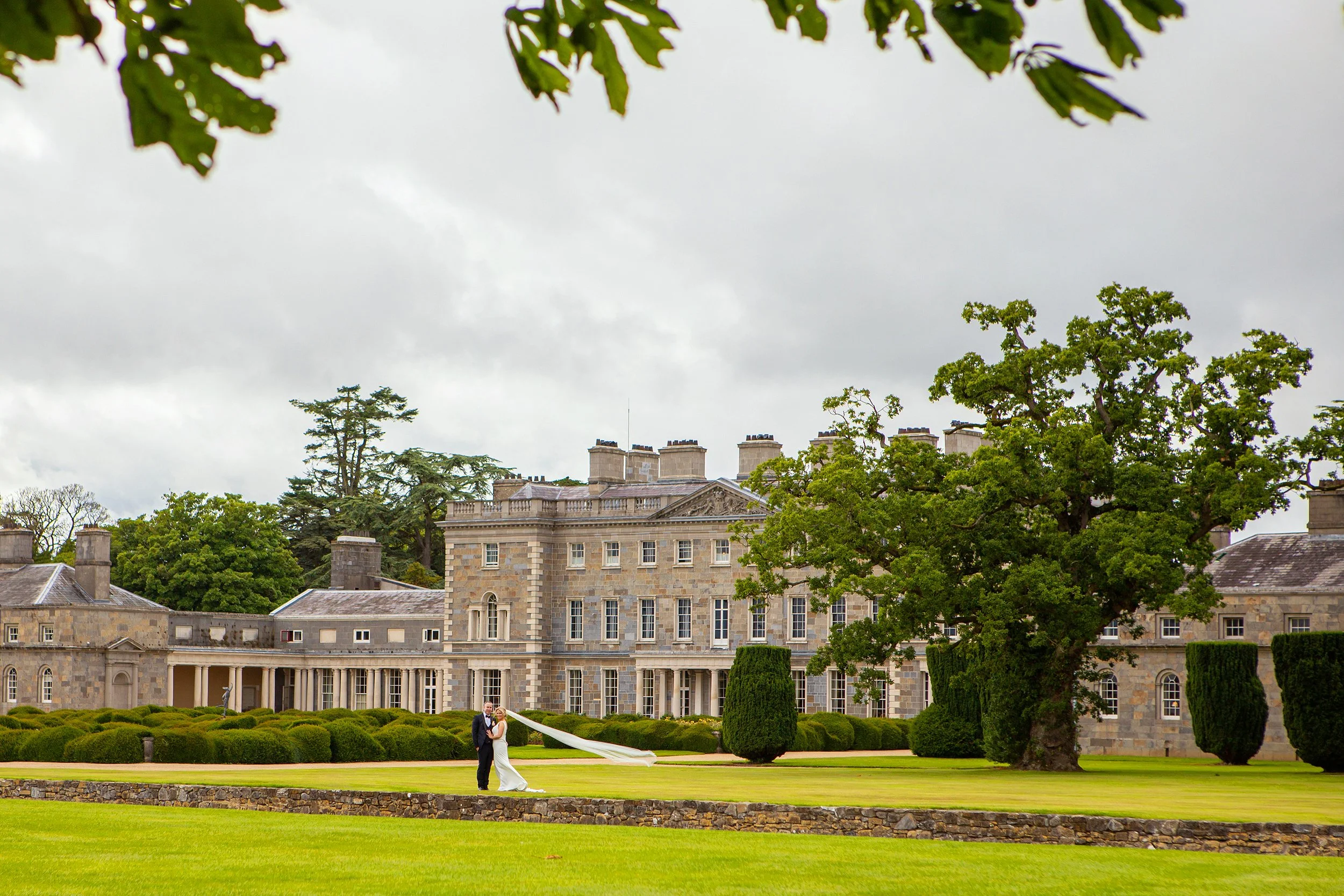 A couple dressed in wedding attire stands on a grassy lawn in front of a large stone building, with trees and shrubbery surrounding a formal garden area, and a cloudy sky overhead.