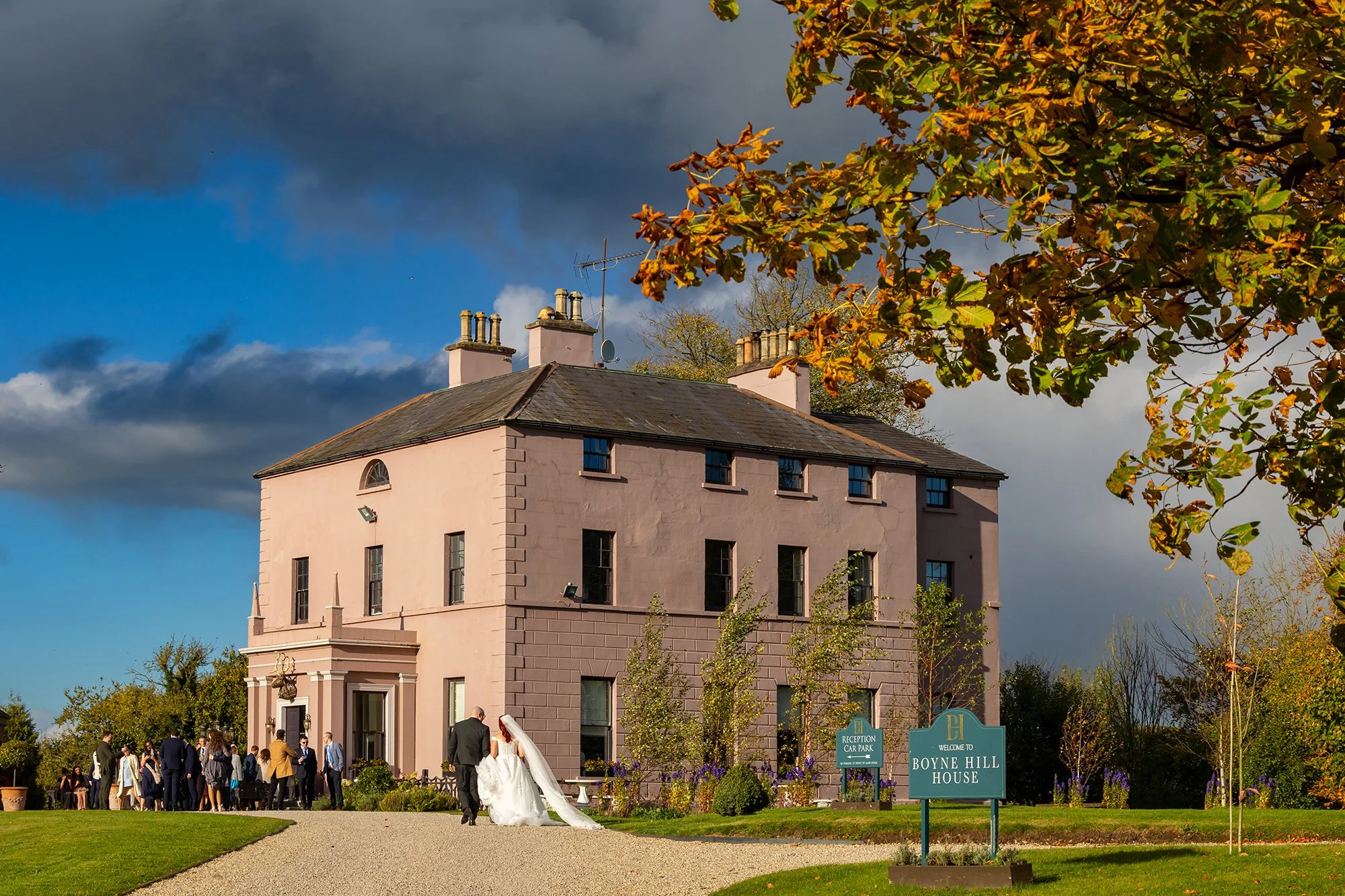 A wedding party gathering outside a pink historic house called Boyne Hill House with people mingling on the lawn and a bride in a white wedding gown walking with a person in a suit under a tree with autumn-colored leaves. Dark storm clouds are in the sky.