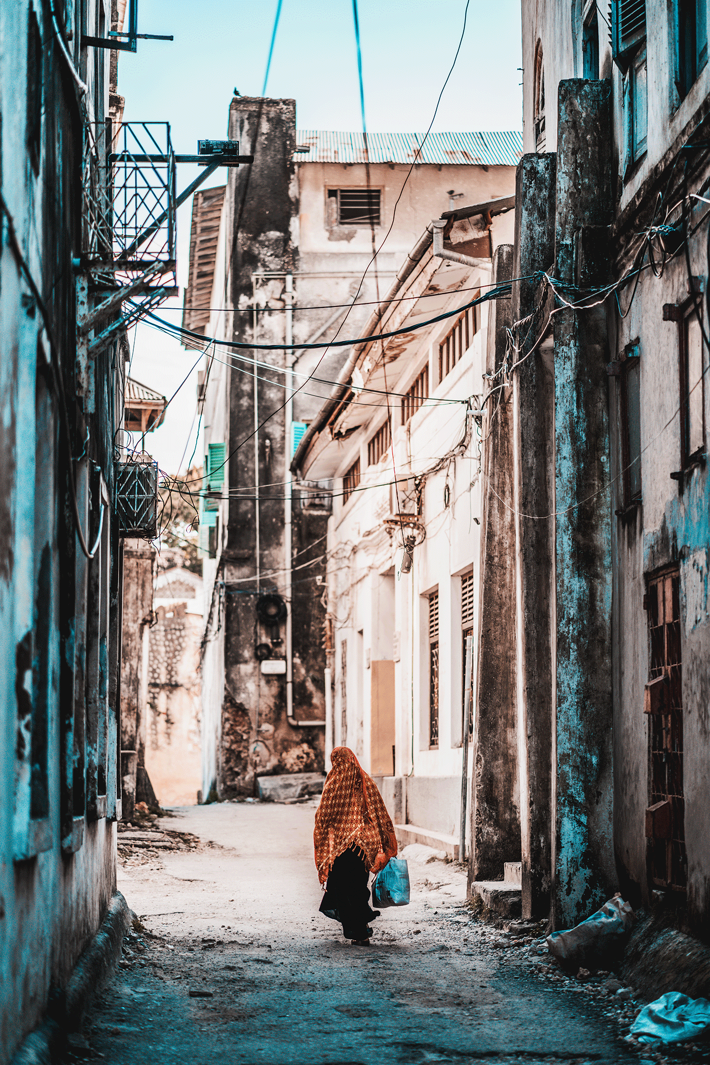 A young girl walking down a narrow, dirt alleyway with worn and weathered buildings on either side, carrying a blue bag and wearing a patterned headscarf.