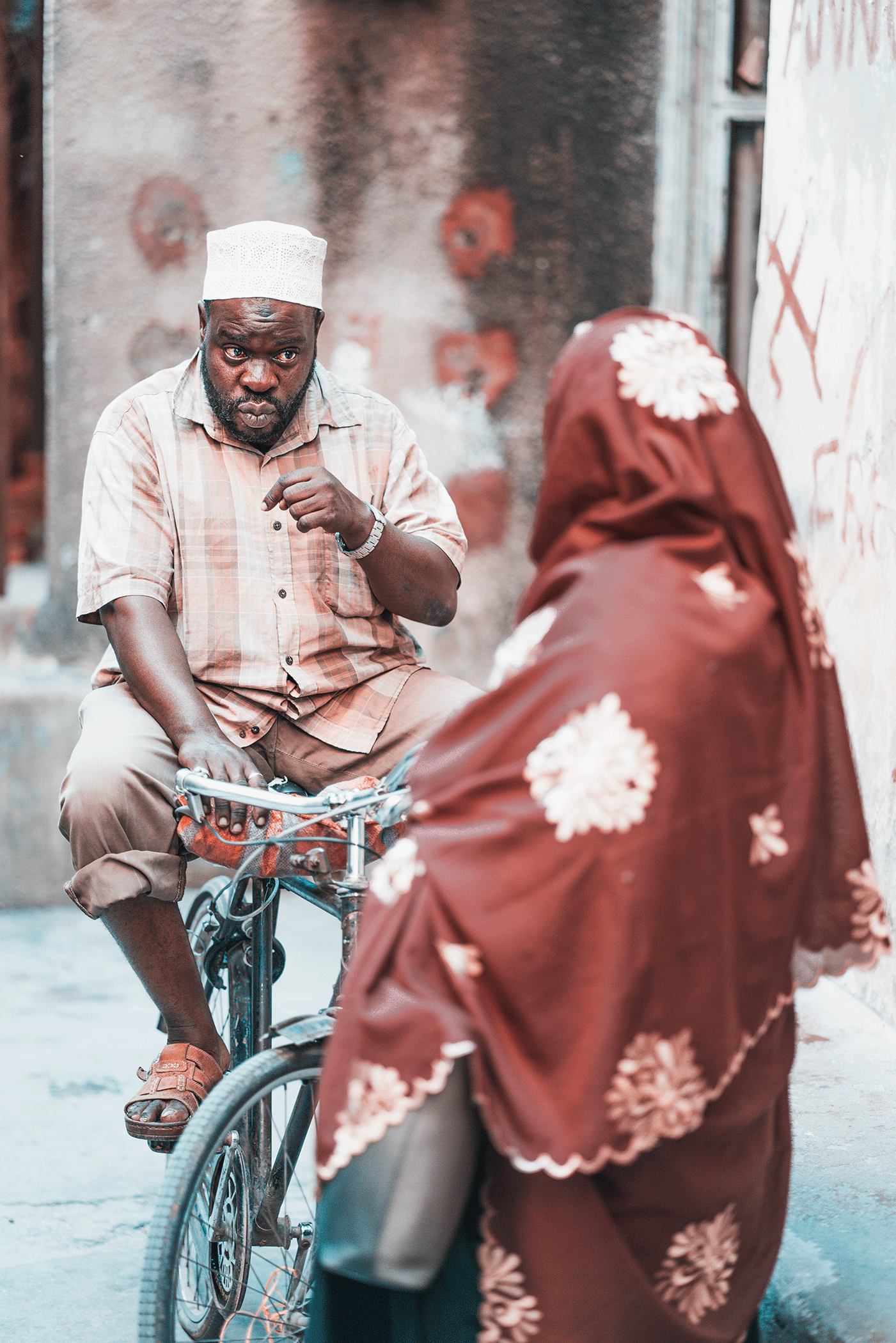 An older man with a beard and wearing a white hat is sitting on a bicycle, looking serious as he talks to a woman in a brown headscarf with white floral patterns, who has her back to the camera, against a textured wall with graffiti.