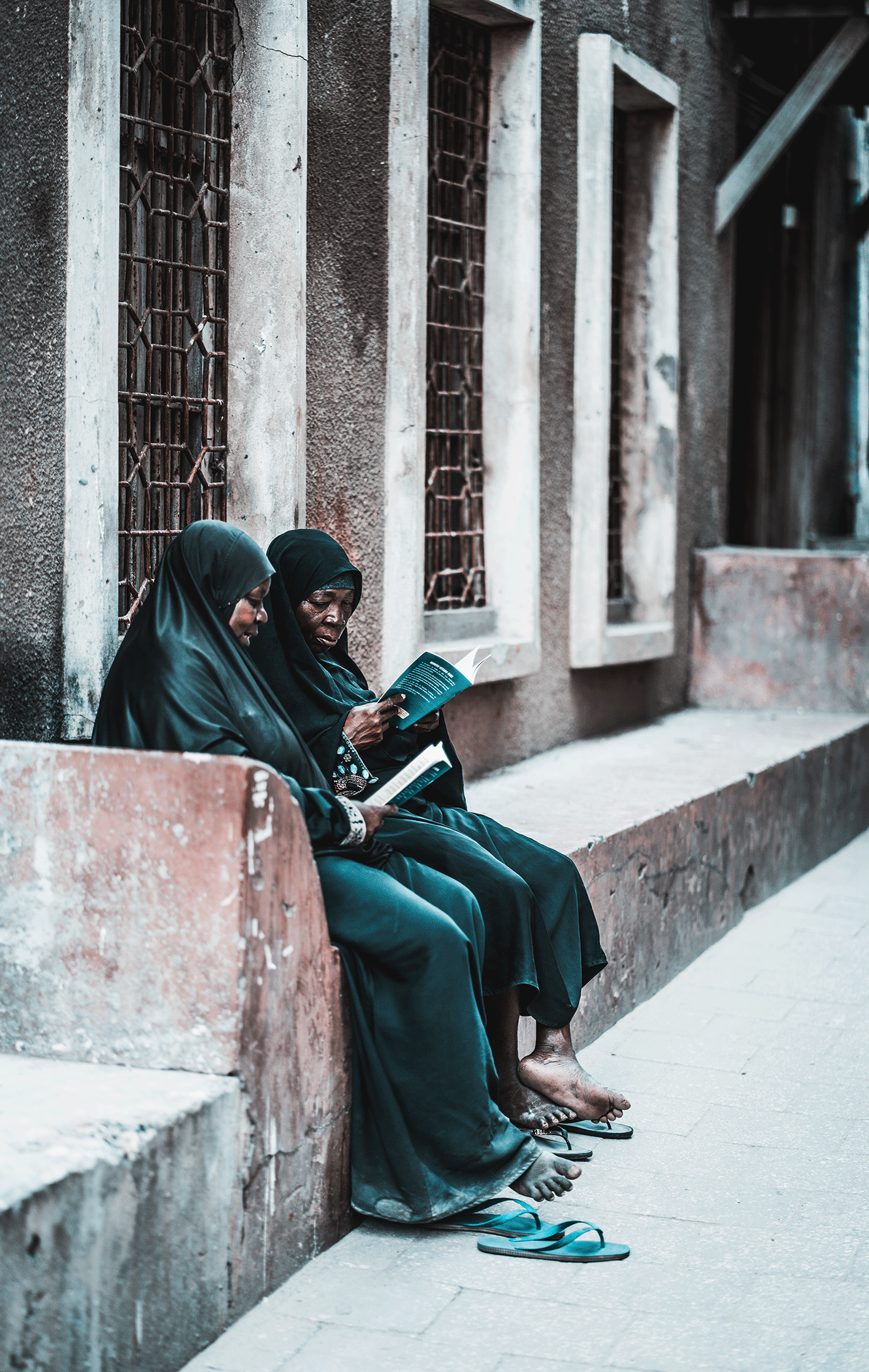 Two elderly women sitting on a concrete wall reading books, wearing black headscarves and black clothing, with sandals and slippers on the ground in front of them, next to a building with barred windows.
