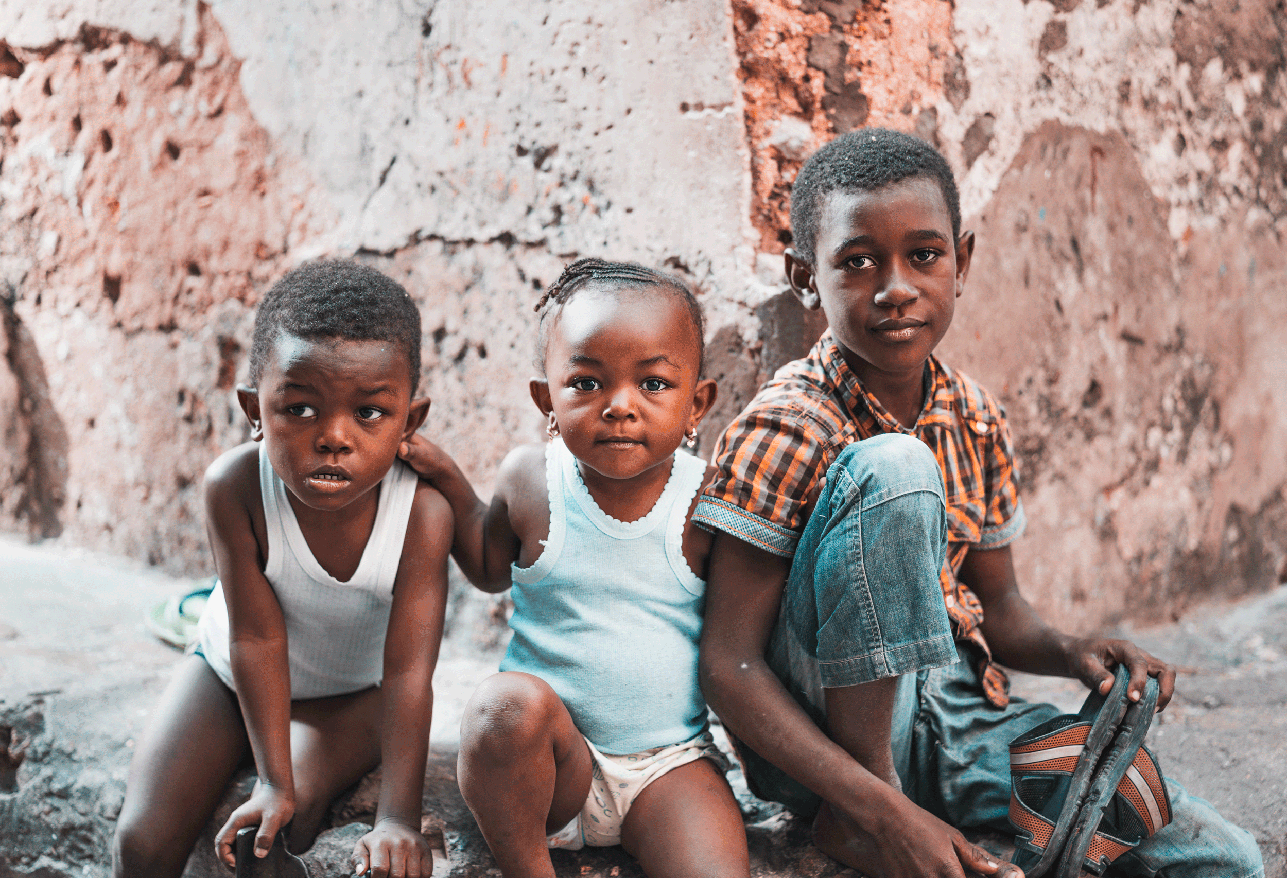 Three children sitting against a rough brick and concrete wall, looking at the camera. The children include two boys and one girl, all with dark skin and short hair, in casual clothing.
