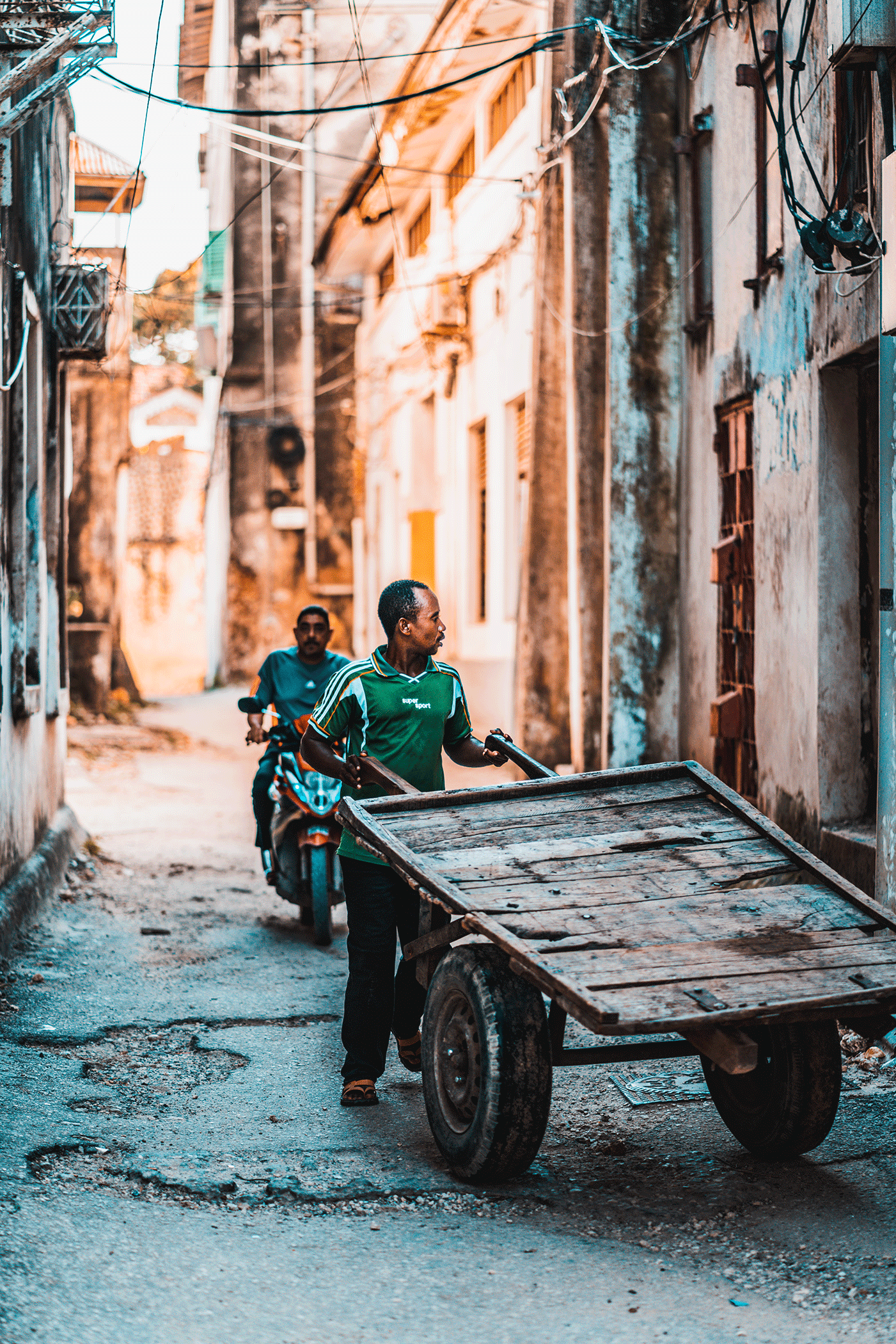 A man pushes a wooden cart down a dirt alley lined with worn buildings, with another man riding a motorcycle behind him.