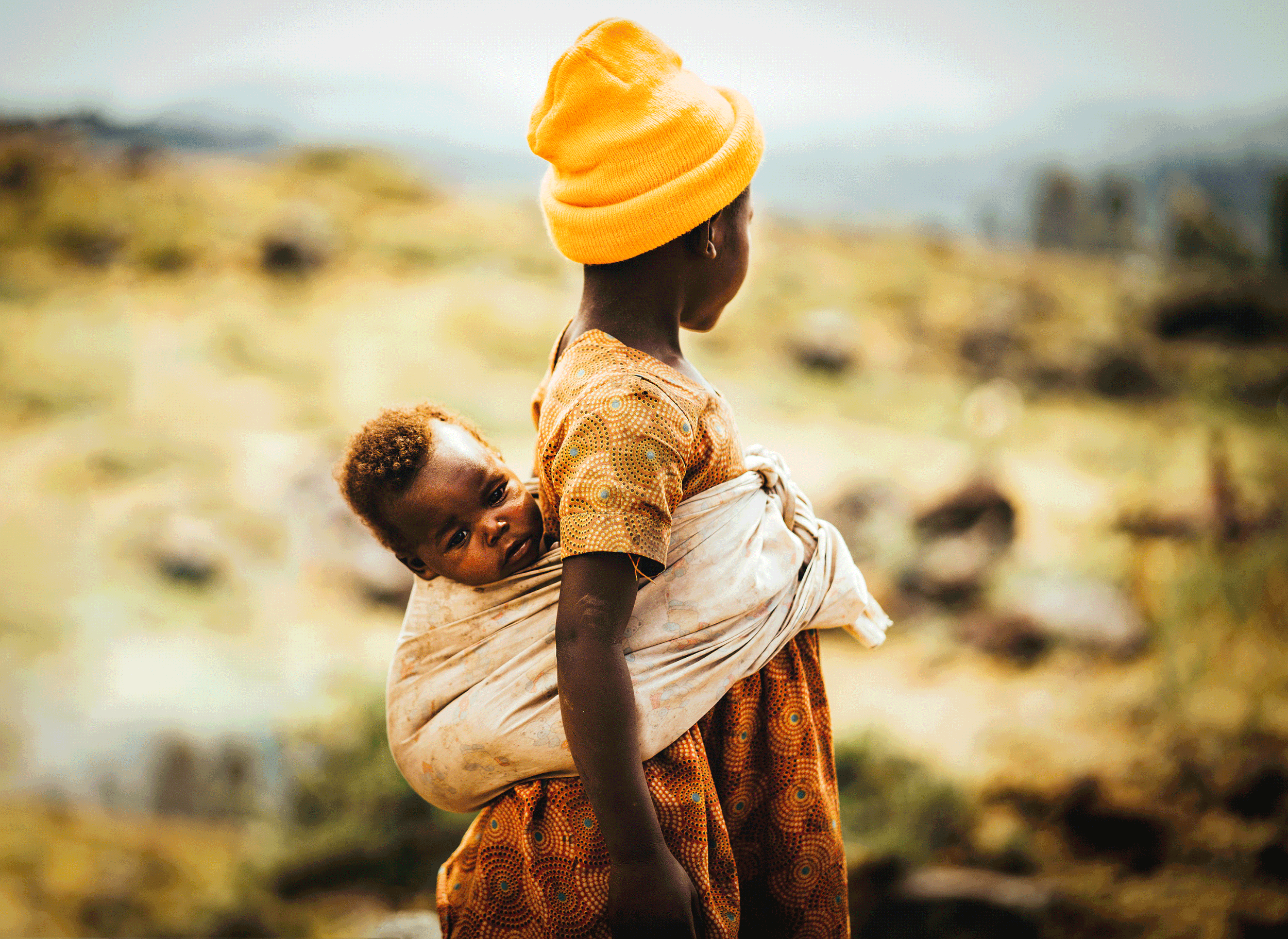 A woman carrying a child on her back outdoors. Both are dressed in traditional clothing, with the woman wearing a bright yellow hat and patterned dress, and the child wrapped in a cloth.