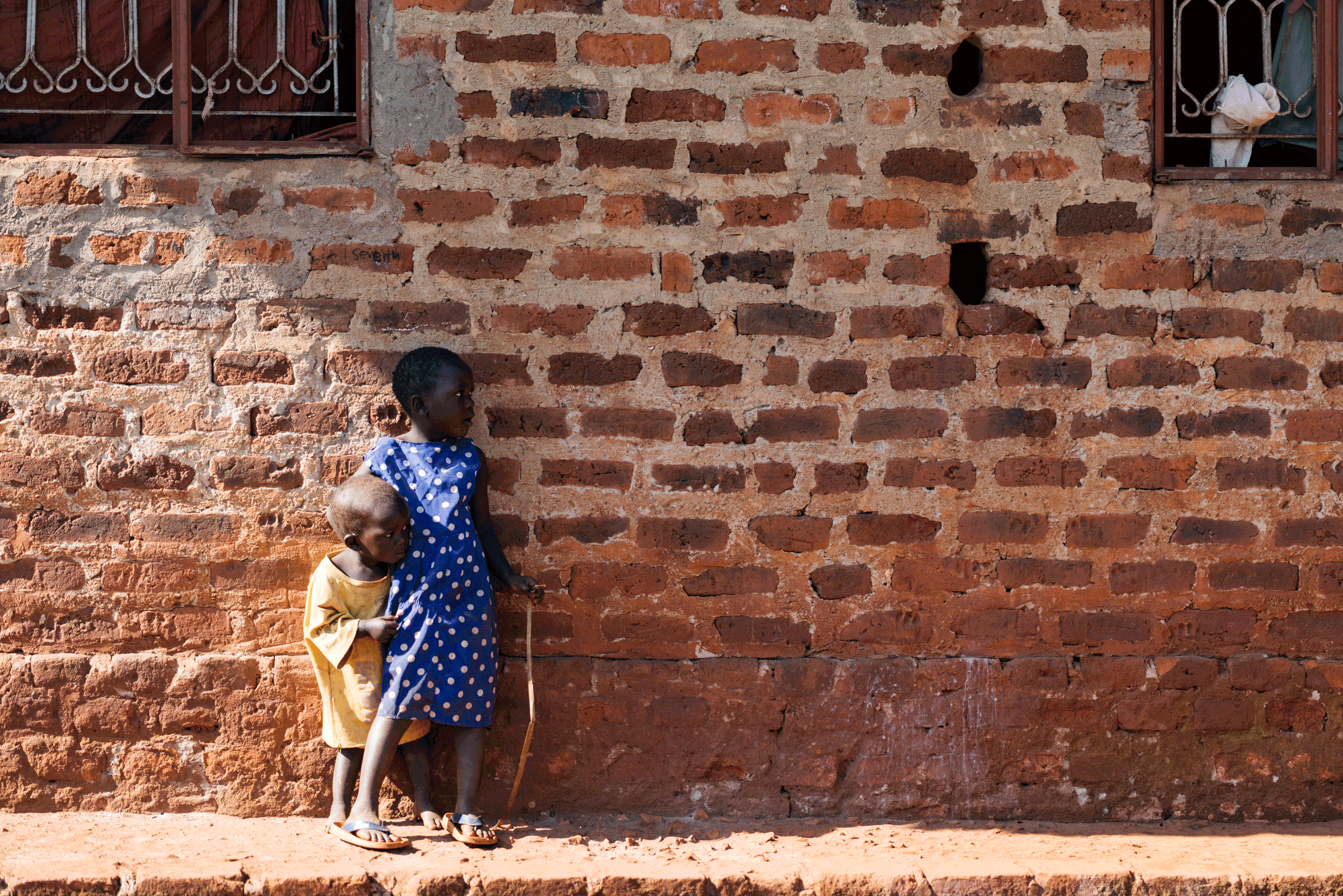 Two young children standing against a red brick wall, one girl holding a stick, both children wearing dresses and sandals.