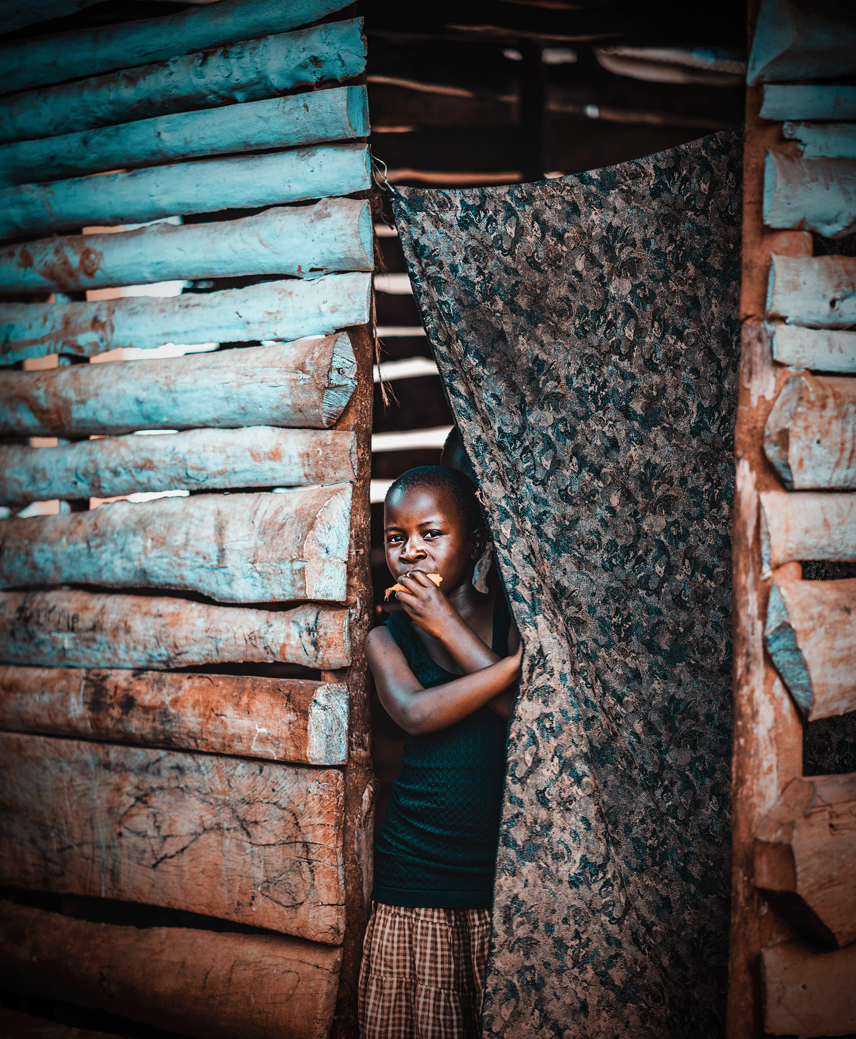 A young boy peeking out from behind a makeshift wall made of stacked logs, holding a piece of bread and looking directly at the camera.