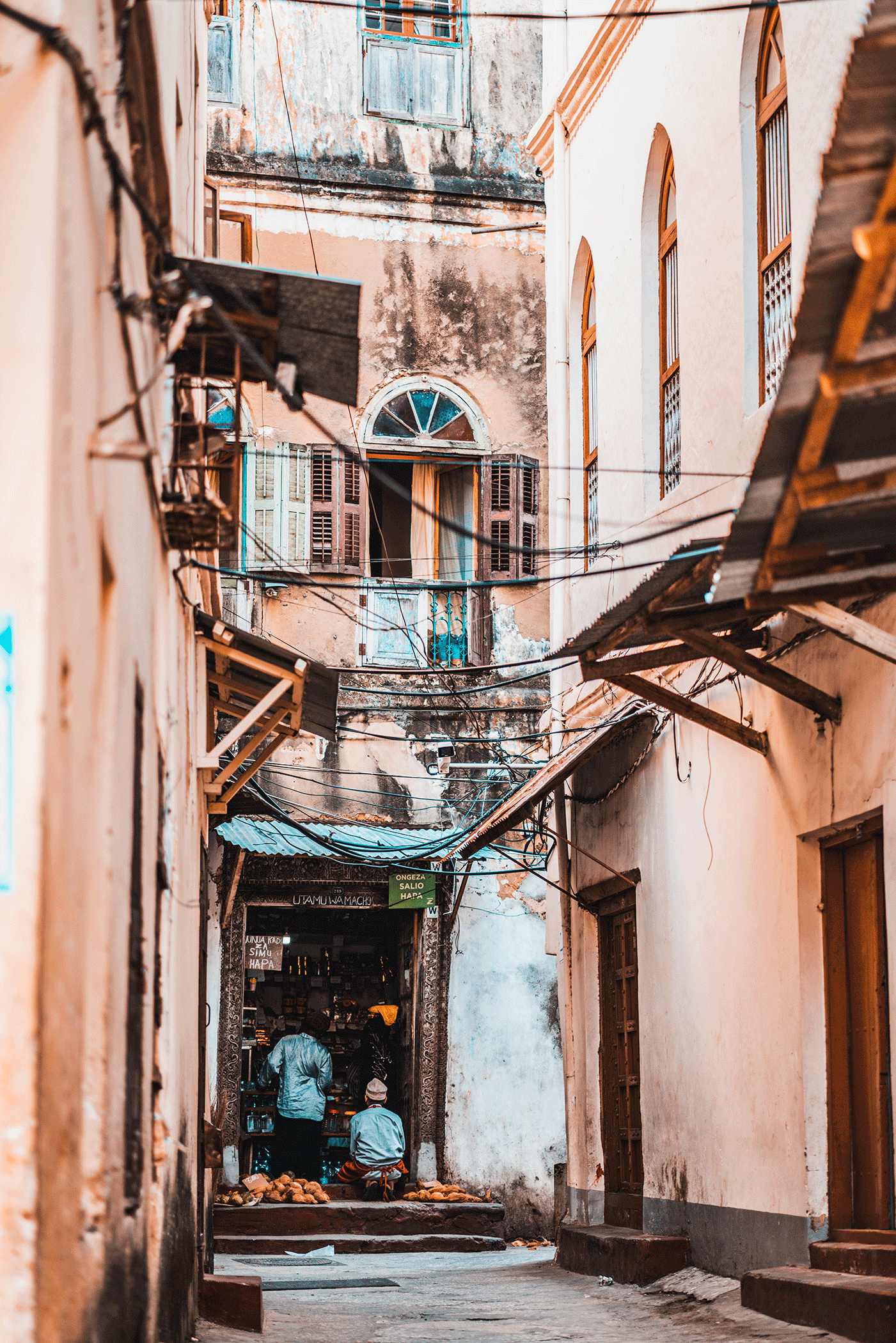 Narrow street in an old city with worn, colorful buildings and open windows. Two men are walking towards a shop entrance surrounded by black ornate decorative archway, with several bread baguettes on the ground.