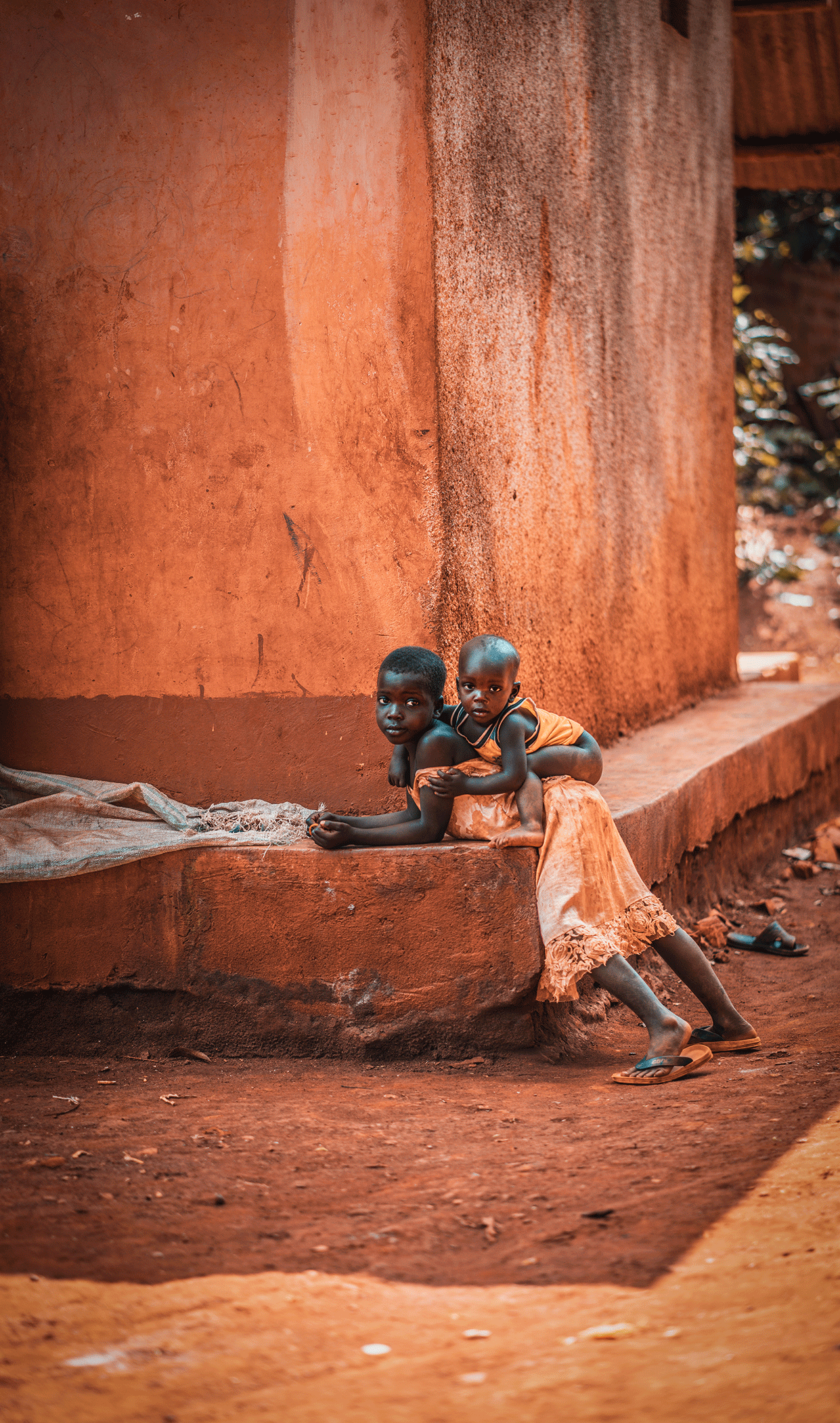Two young children with dark skin sitting on a concrete ledge in front of a large reddish-brown wall. One child is lying down while the other is sitting behind, with their arms around each other. The children are dressed in simple clothing, and there