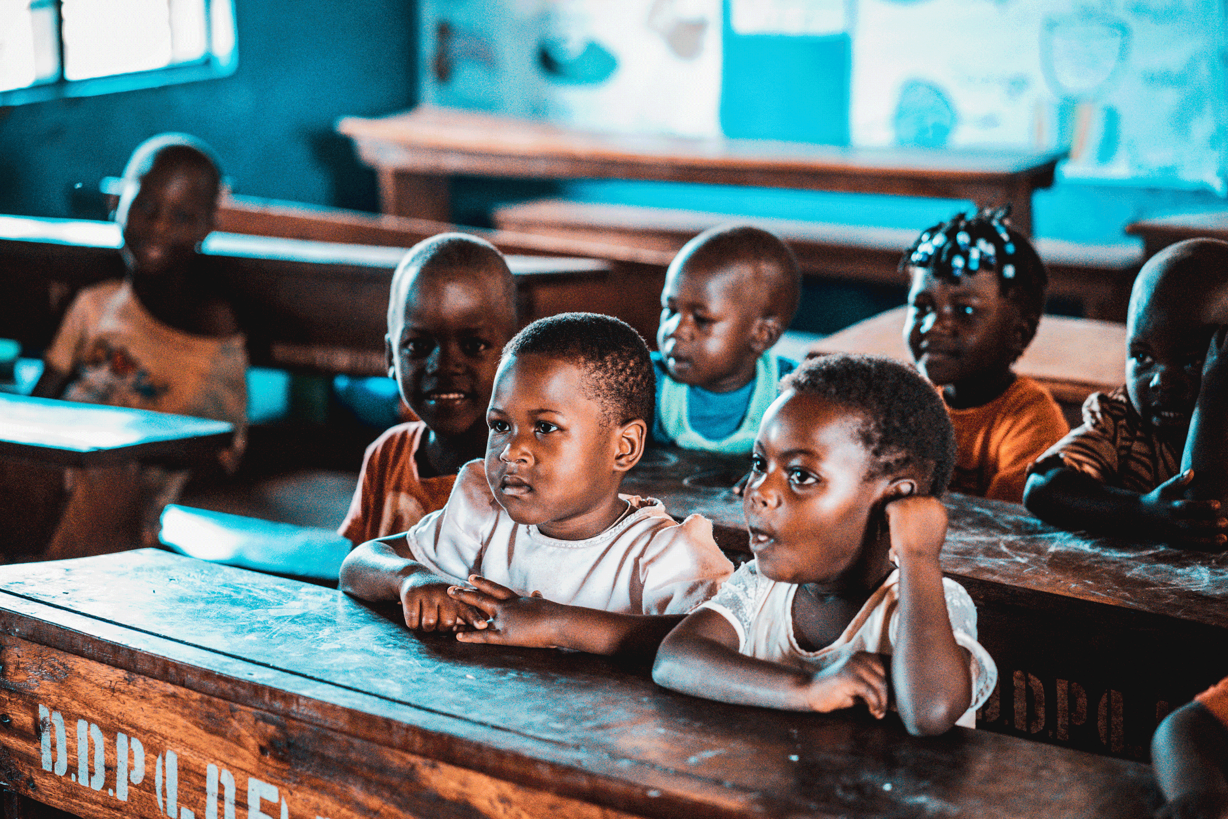 A classroom with young children sitting at wooden desks, attentively facing forward, some with serious expressions and others slightly smiling.