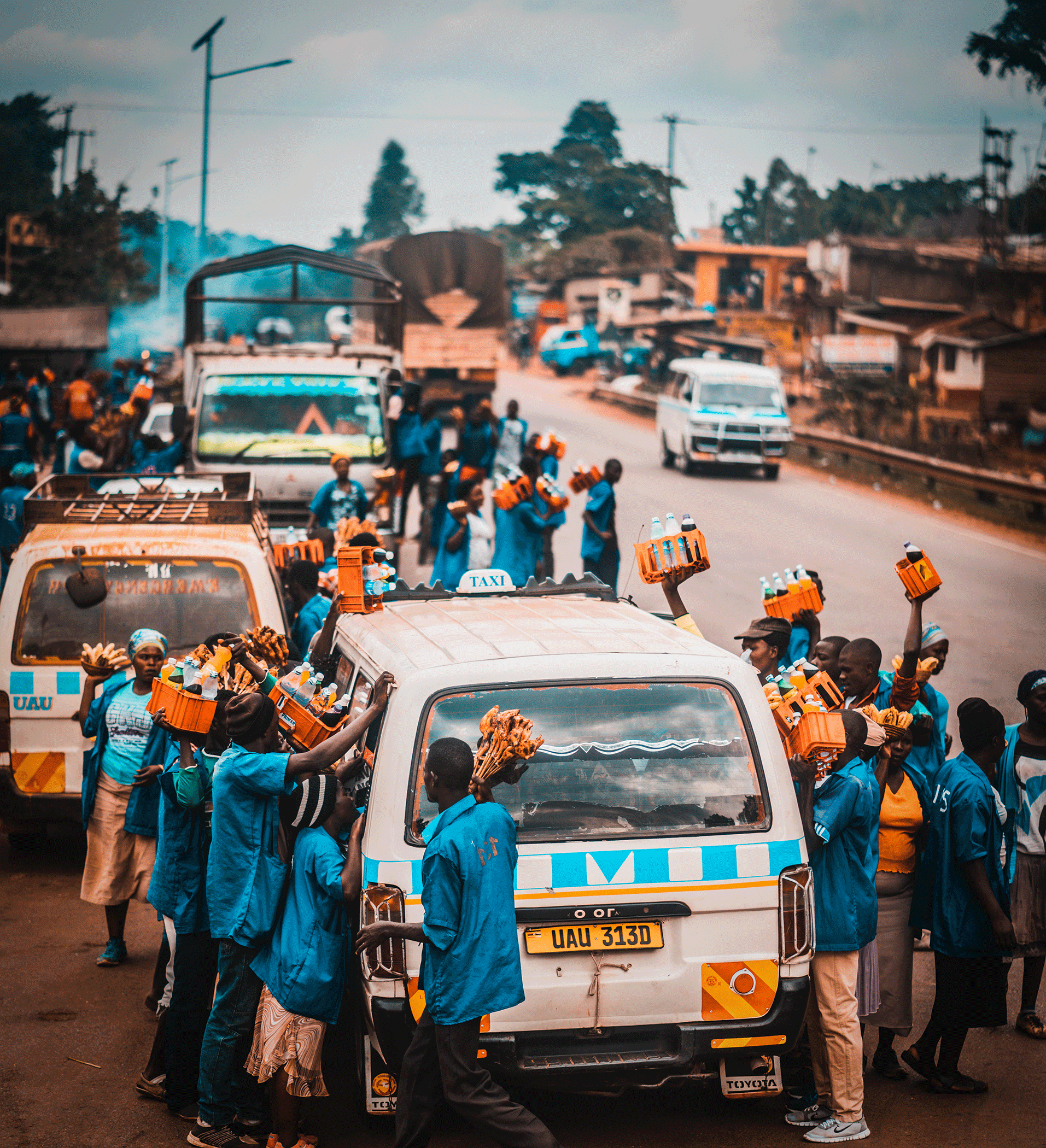 A street scene in an African town with people walking and stacking orange crates filled with bottles on top of a white vehicle with a yellow license plate reading 'U AU 313D.' The vehicle has a taxi sign on the roof. There are several buildings and t