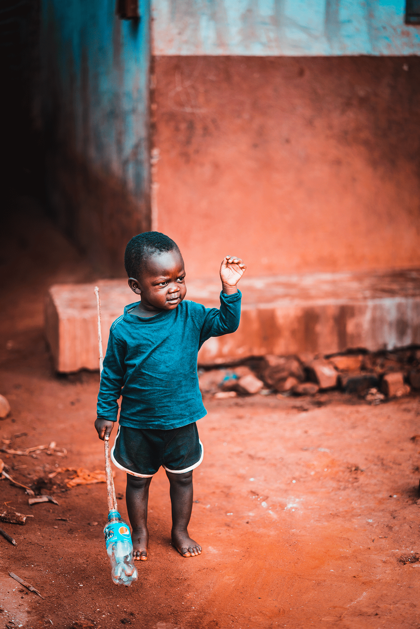 A young boy with dark skin and short hair holding a makeshift spear in one hand and a plastic bottle in the other, standing on a dirt ground in front of a rustic, weathered building with reddish and blue hues.