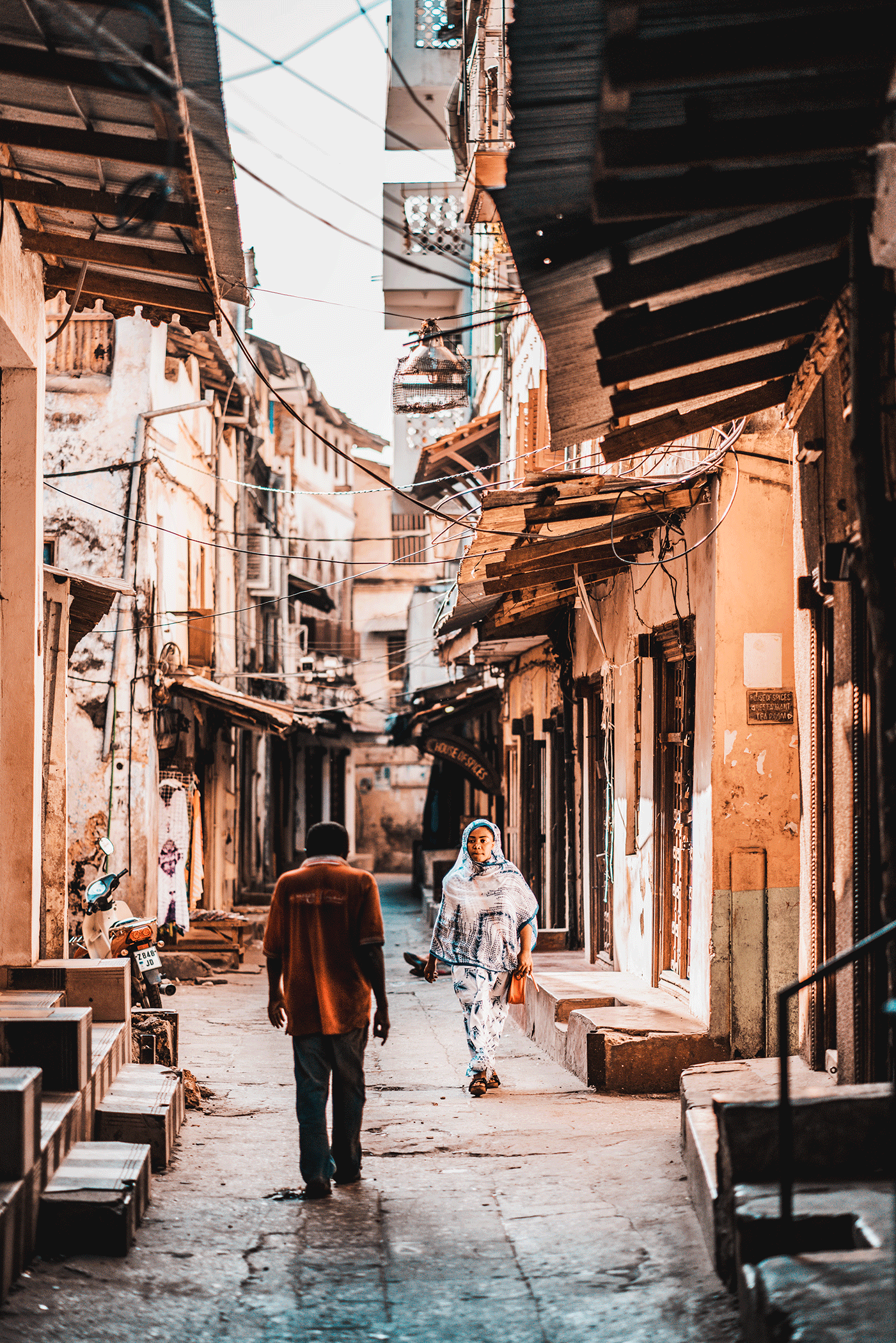 A narrow street in a possibly developing area with old buildings, wires, and street vendors. Two people are walking, one woman wearing a headscarf and a man in a brown shirt.