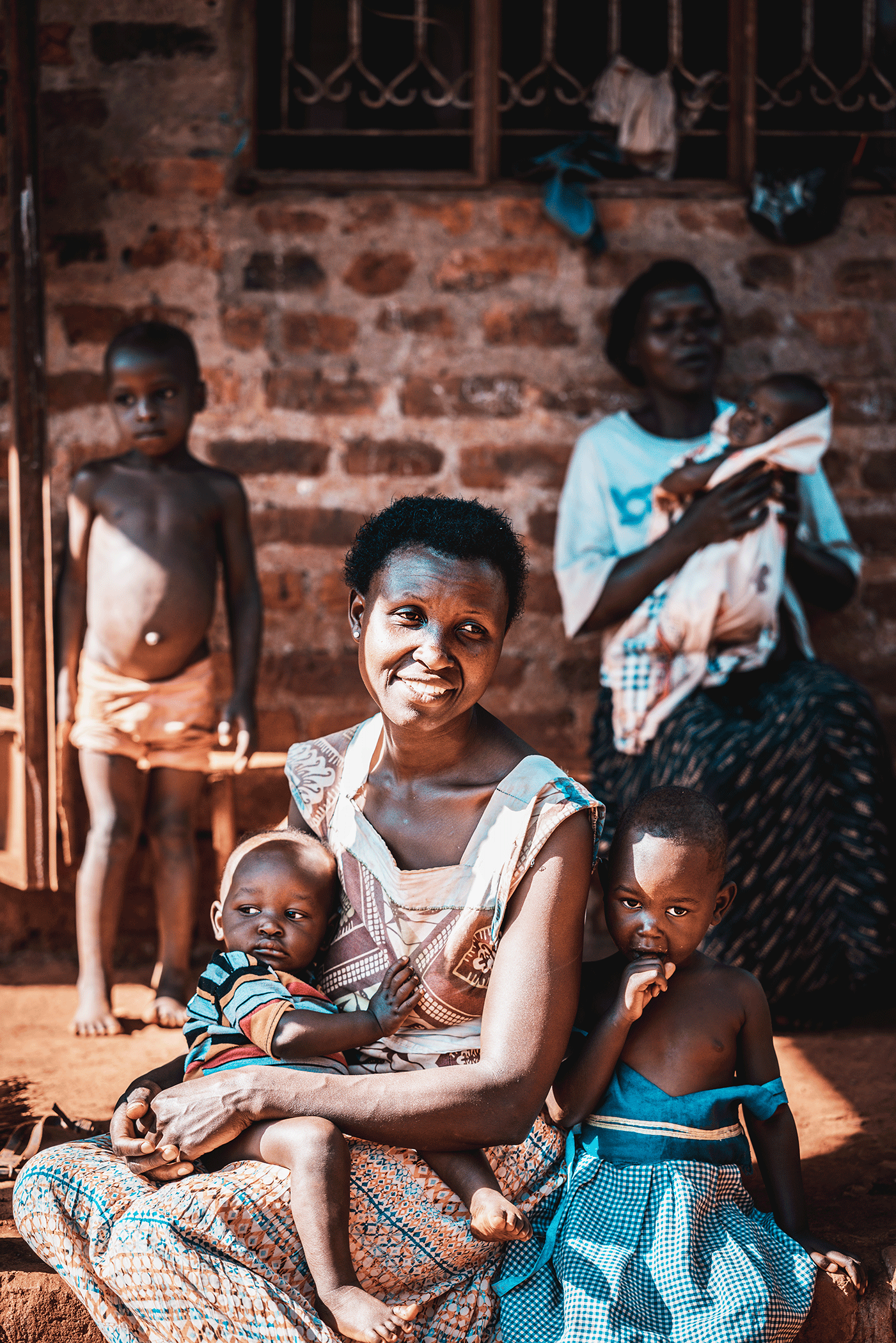 A group of African children and women sitting and standing outside in front of a brick wall, some children are barefoot and wearing minimal clothing, with a woman holding a baby.