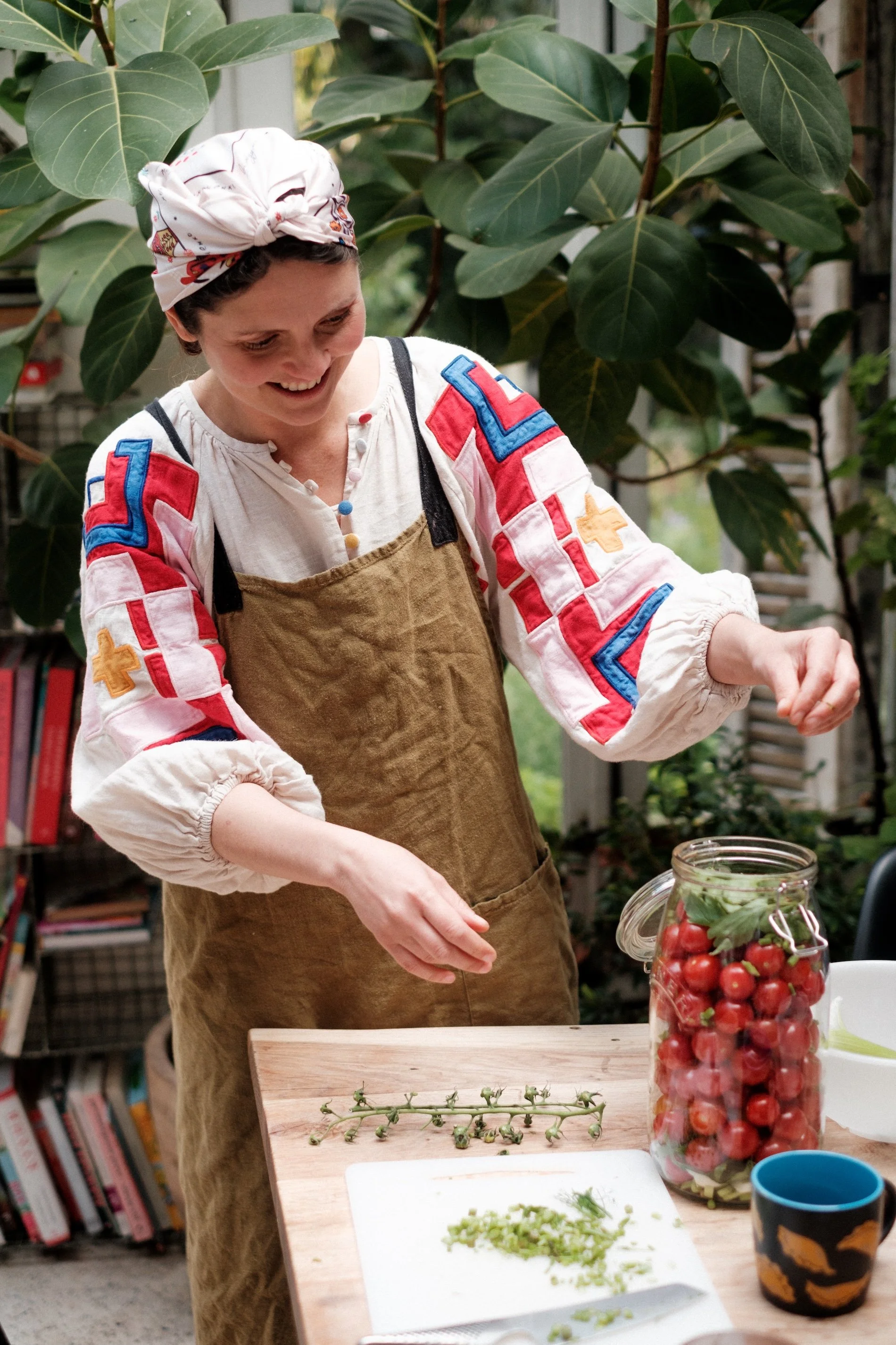 Chef Olia Hercules smiles in font of greenery at she fills a jar with olives to ferment