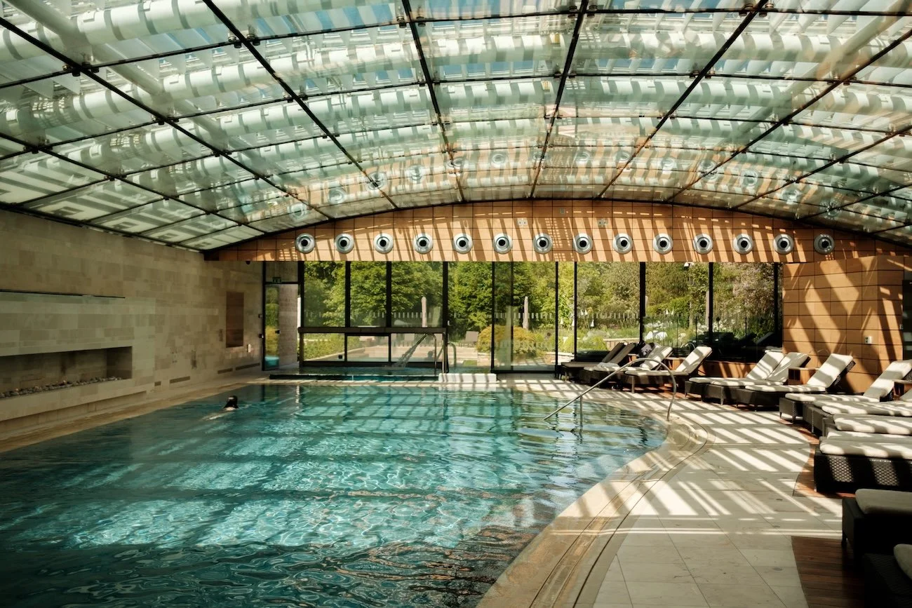 An indoor lap pool is illuminated by sunlight streaming in from the glass ceiling. There is one swimmer and a row of deckchairs