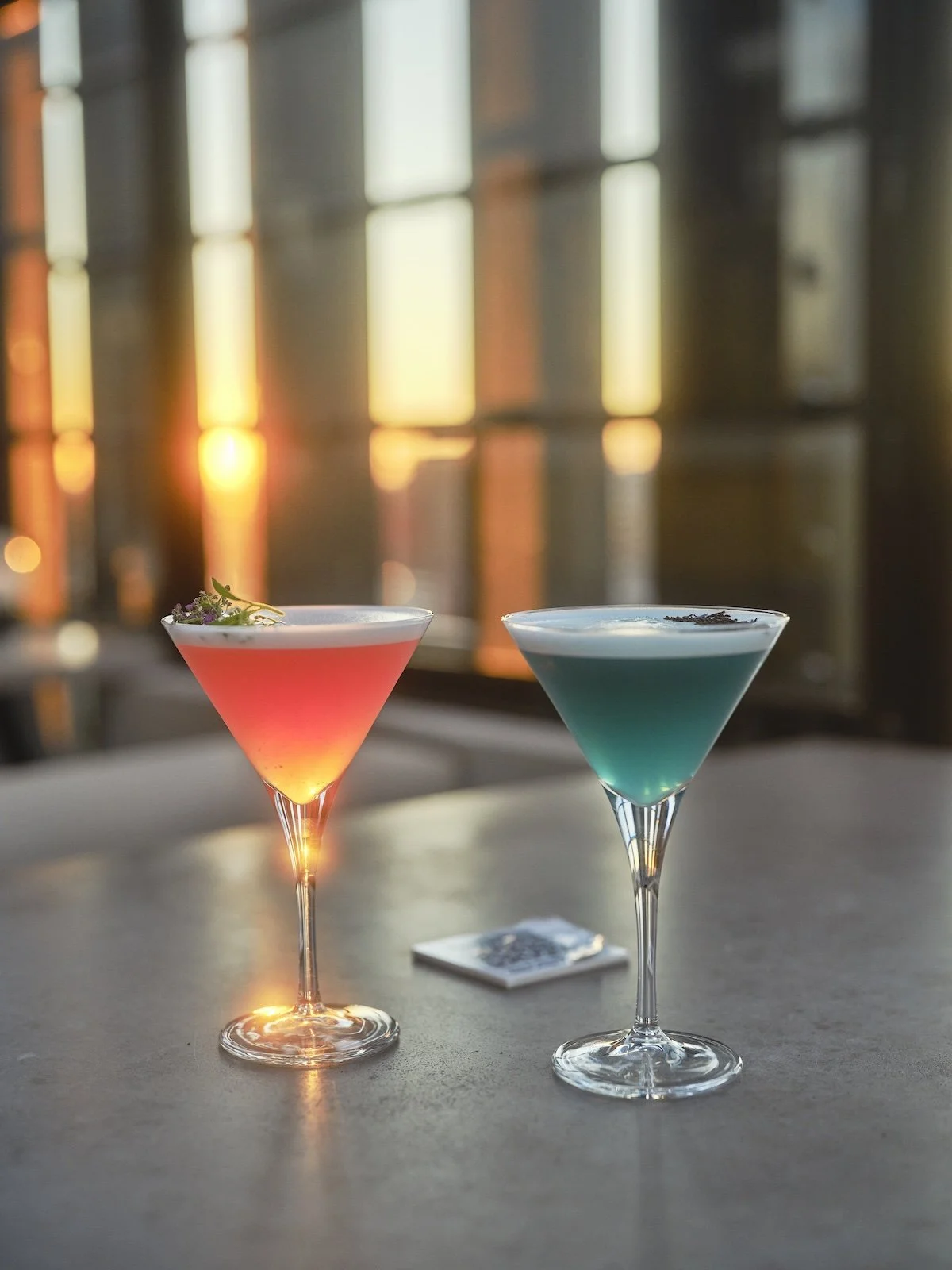 A pink and blue cocktail are illuminated by sunset on a grey outdoor table