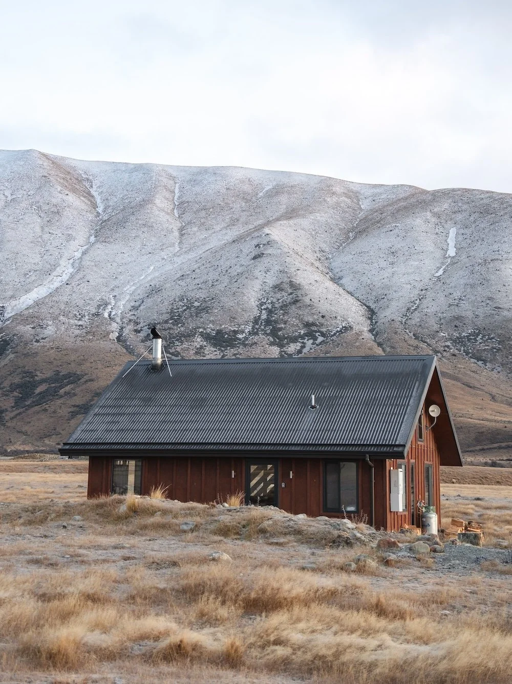 A red cabin with a tin roof sits in a grassy field with a snow covered hill in the background.