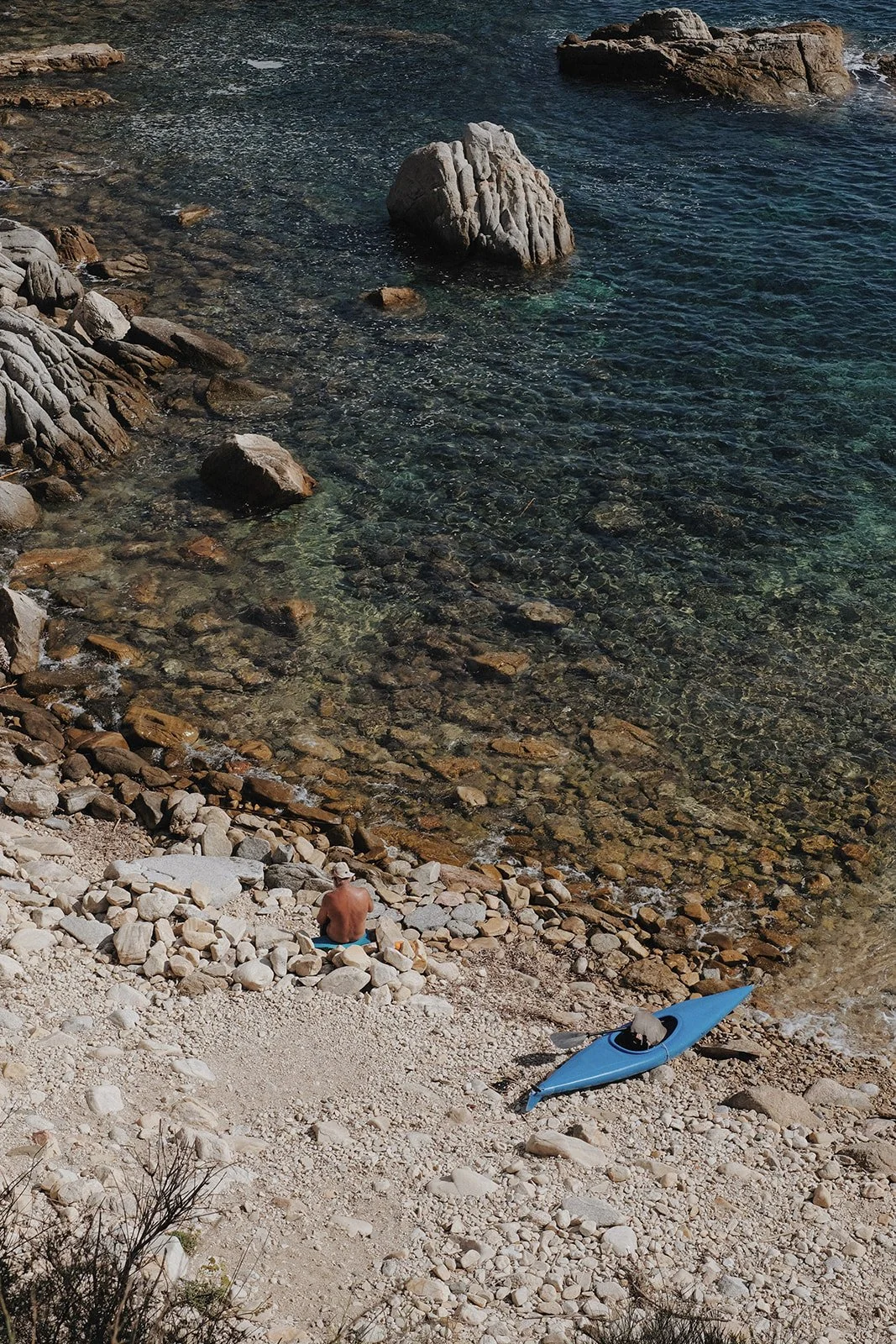 A man sits bu the sea with his blue kayak. The water is so clear that you can see every rock