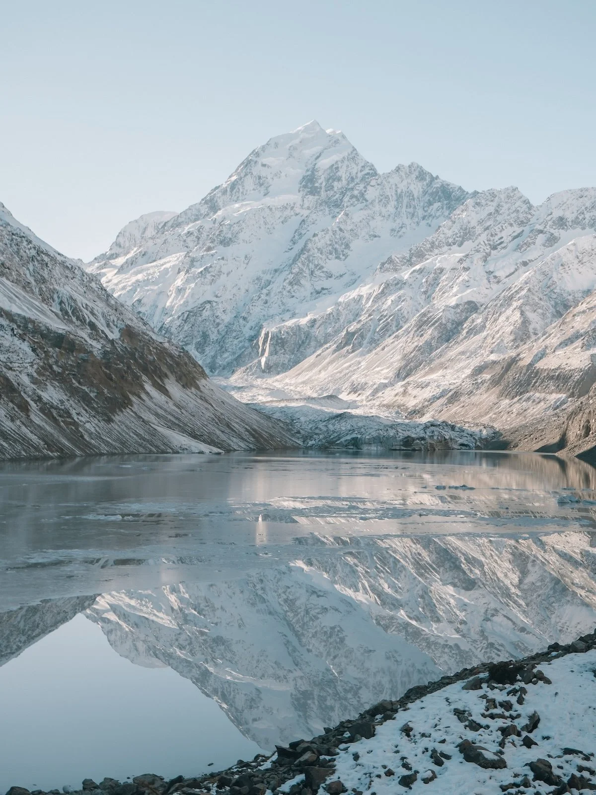 A snow covered mountain, backdropped by blu sky, is reflected in a still alpine lake.