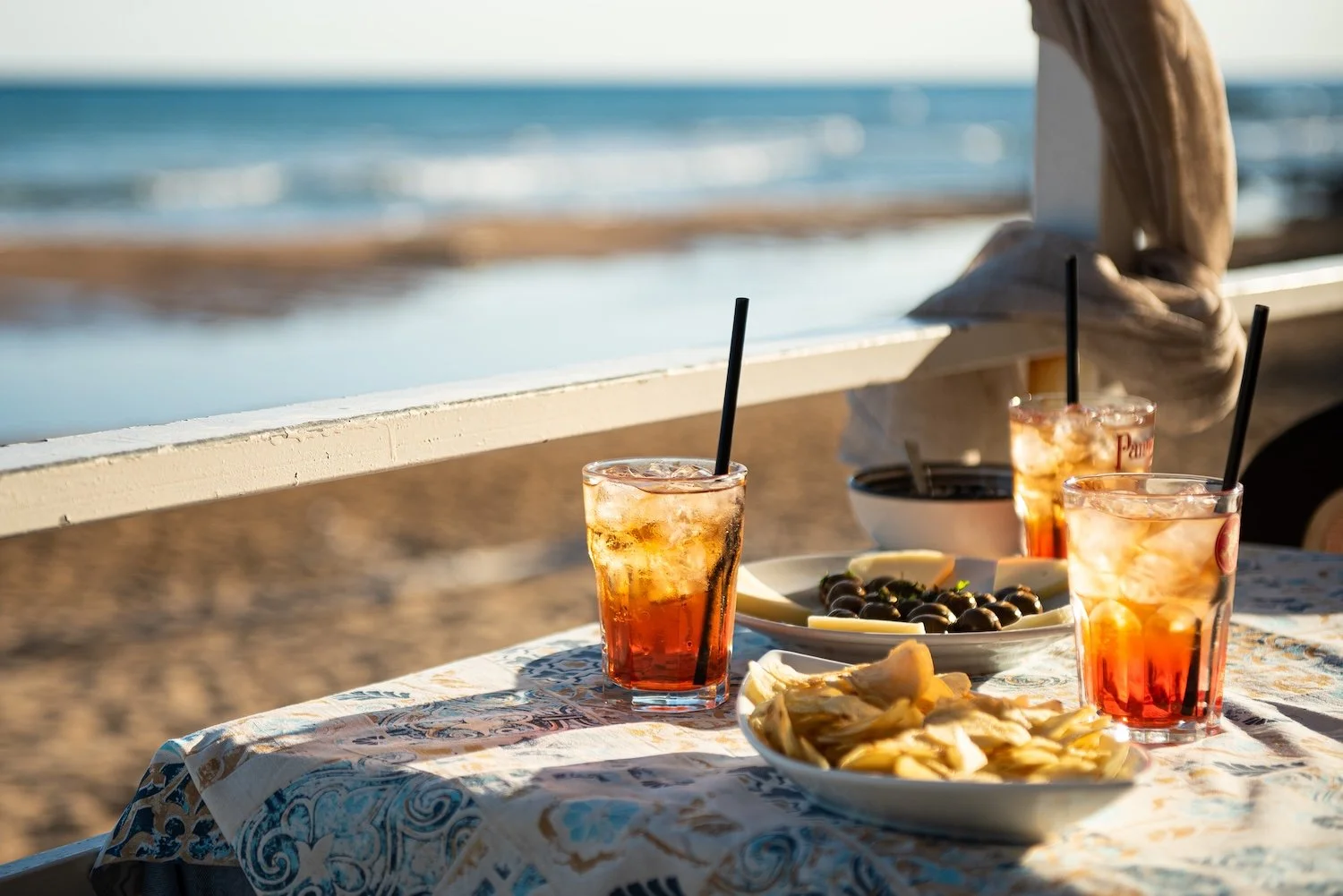 Three glasses of Aperol Spritz sit on a table with the sea visible as a blur in the background