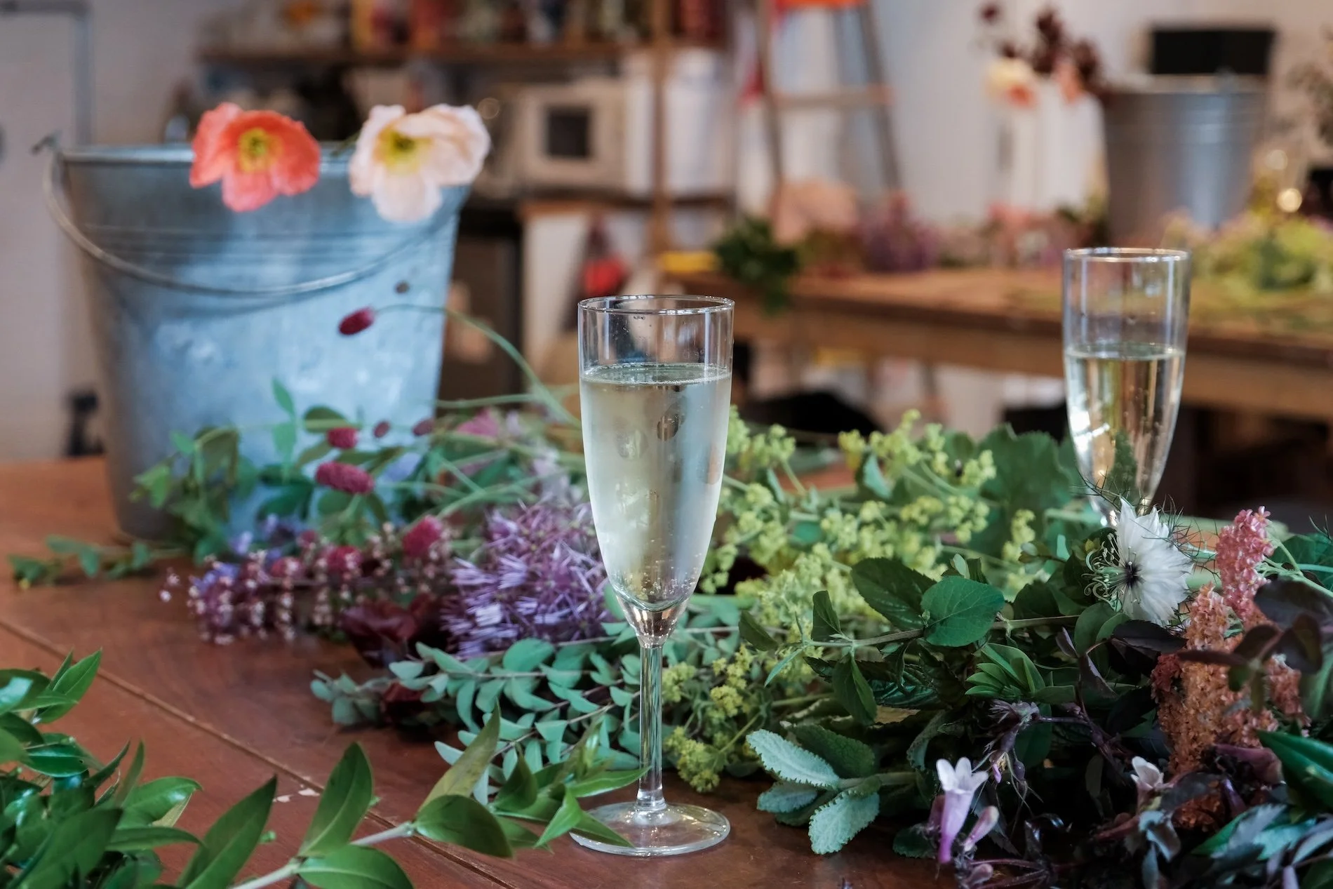 Two glasses of Prosecco sit on a wooden table among flowers ready to be arranged in a workshop