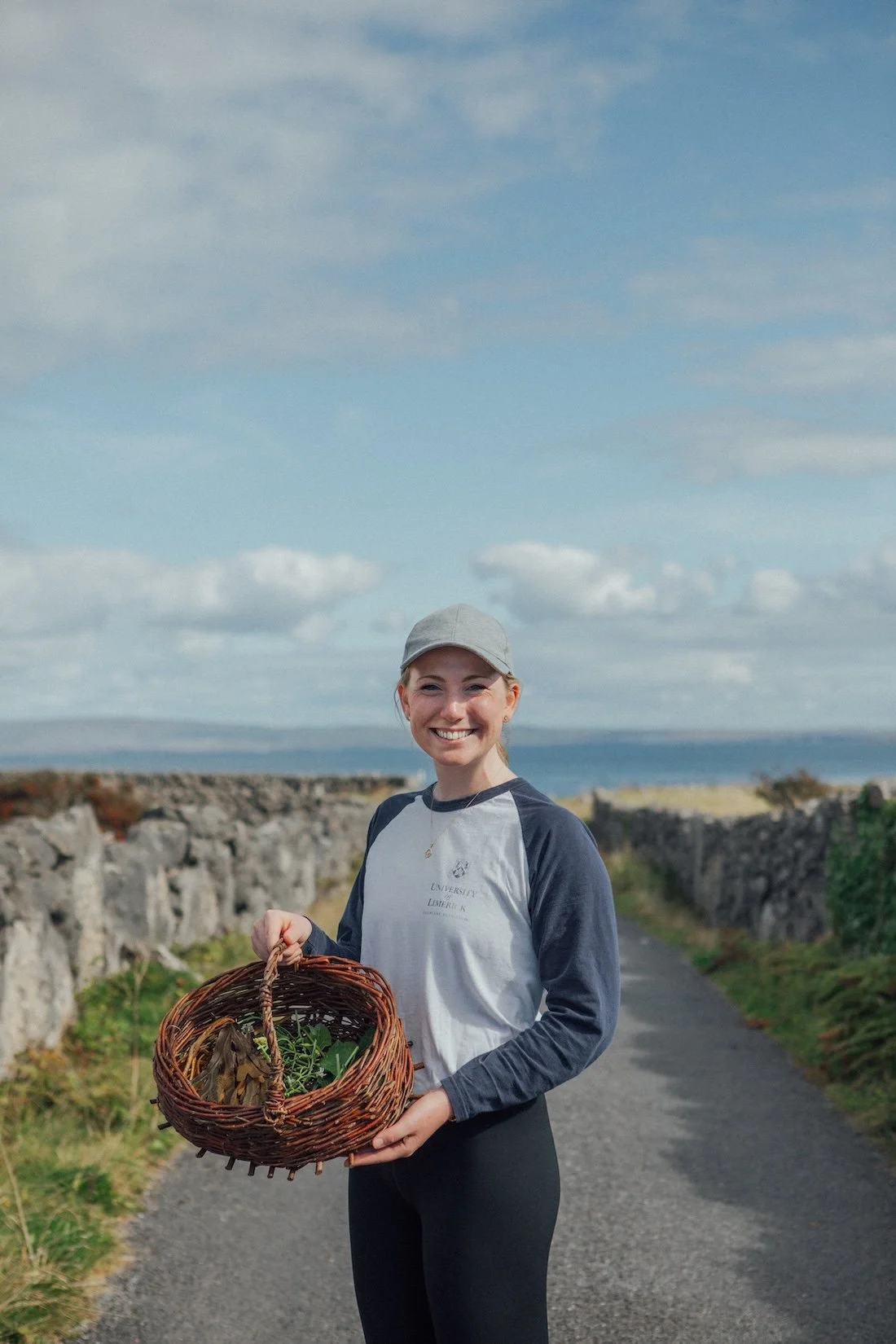 A smiling woman holds her basket of forages herbs and seaweed on a quiet country road with a hint of the ocean behind her