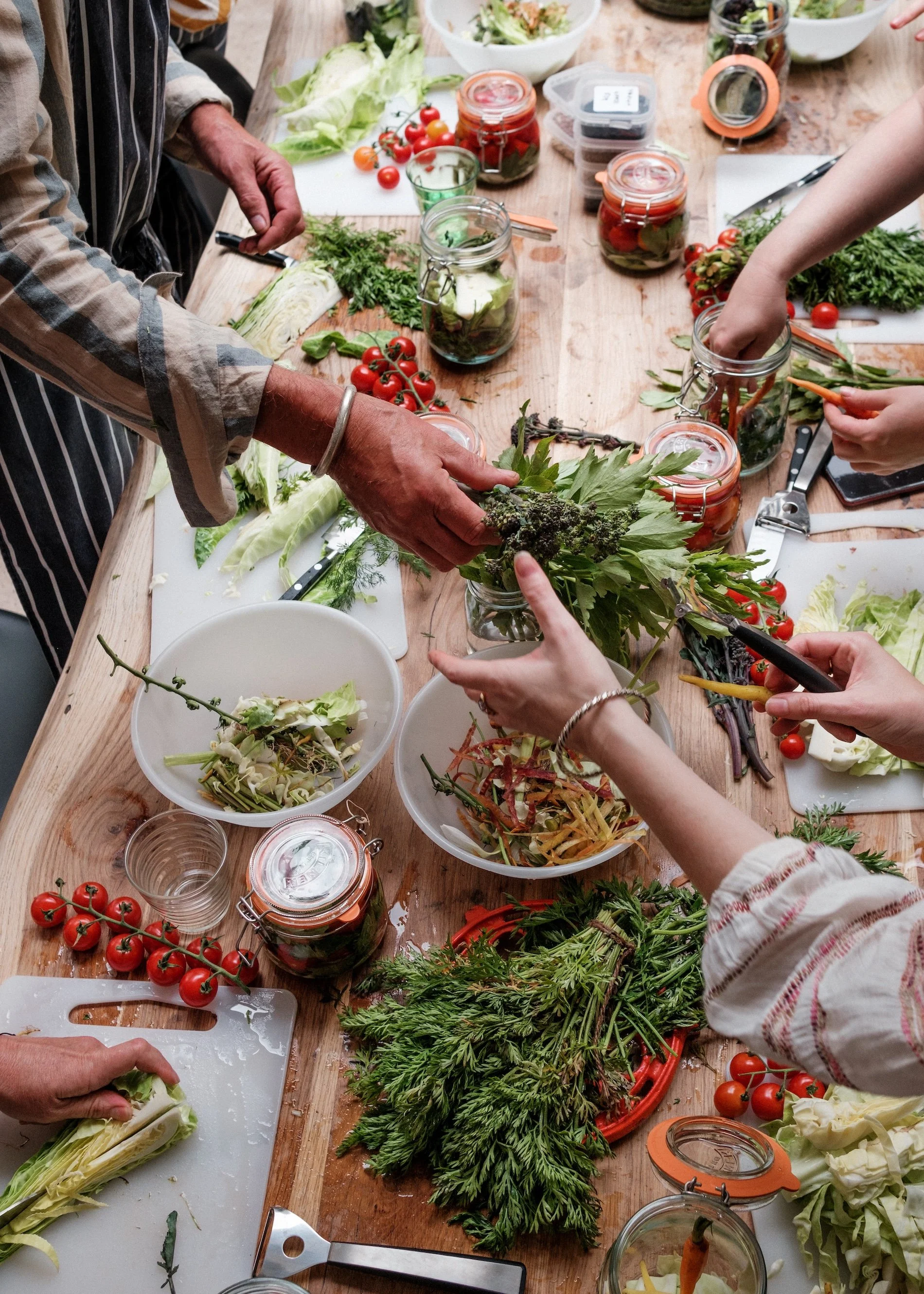 Many hands reach across a wooden table full of vegetables ready to be fermented