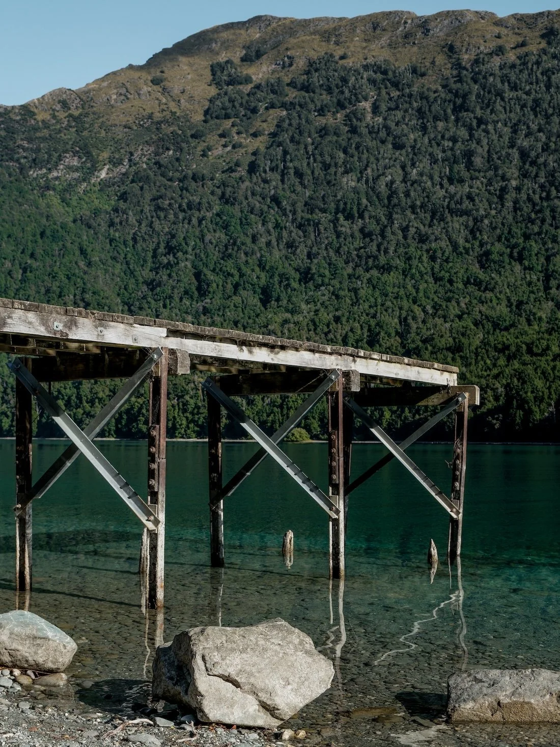 A weathered wooden jetty appears to hover over a lake, which glows green to mirror the tree-covered mountain behind it