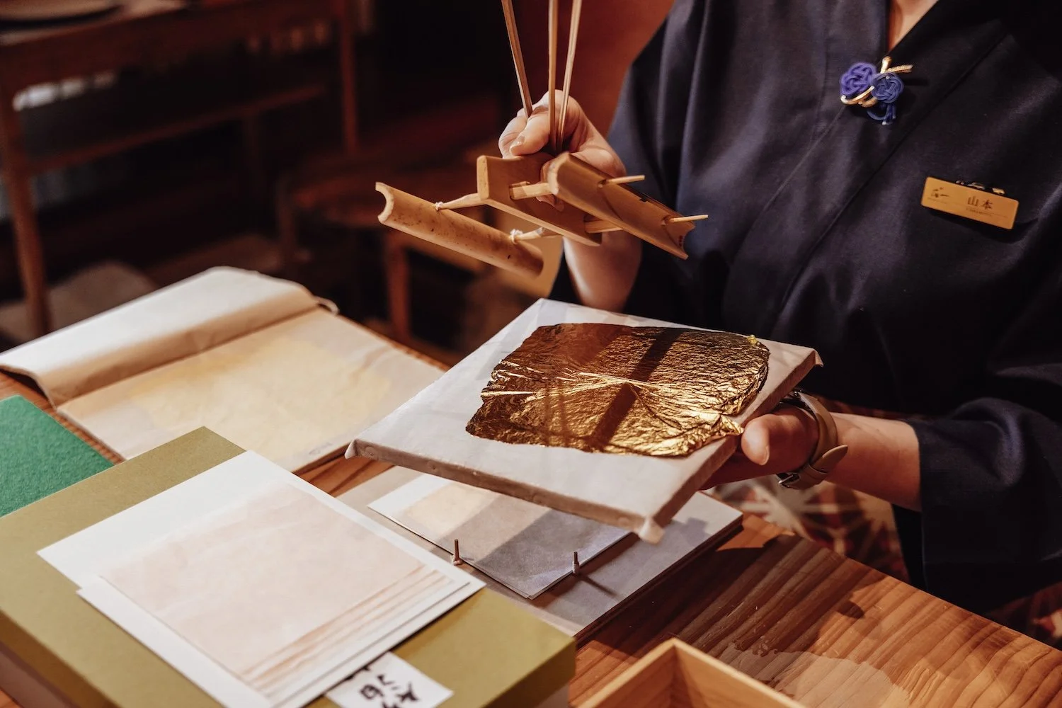 An artisan holds a sheet of gold leaf on a tray. Below her is a table scattered with different types of washi paper