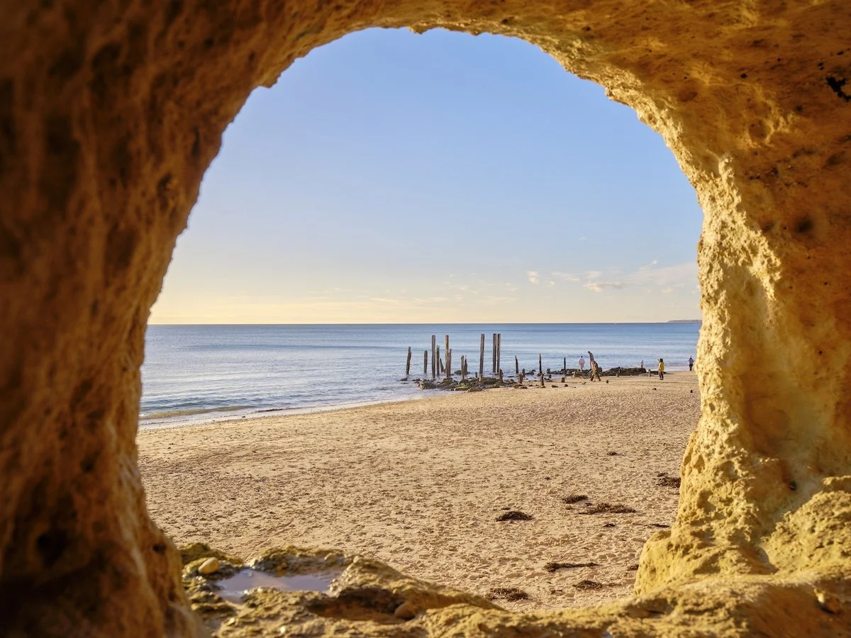 Looking out through the circular opening on a sandstone cave onto the remains of an old wooden jetty rising from a sandy, still beach